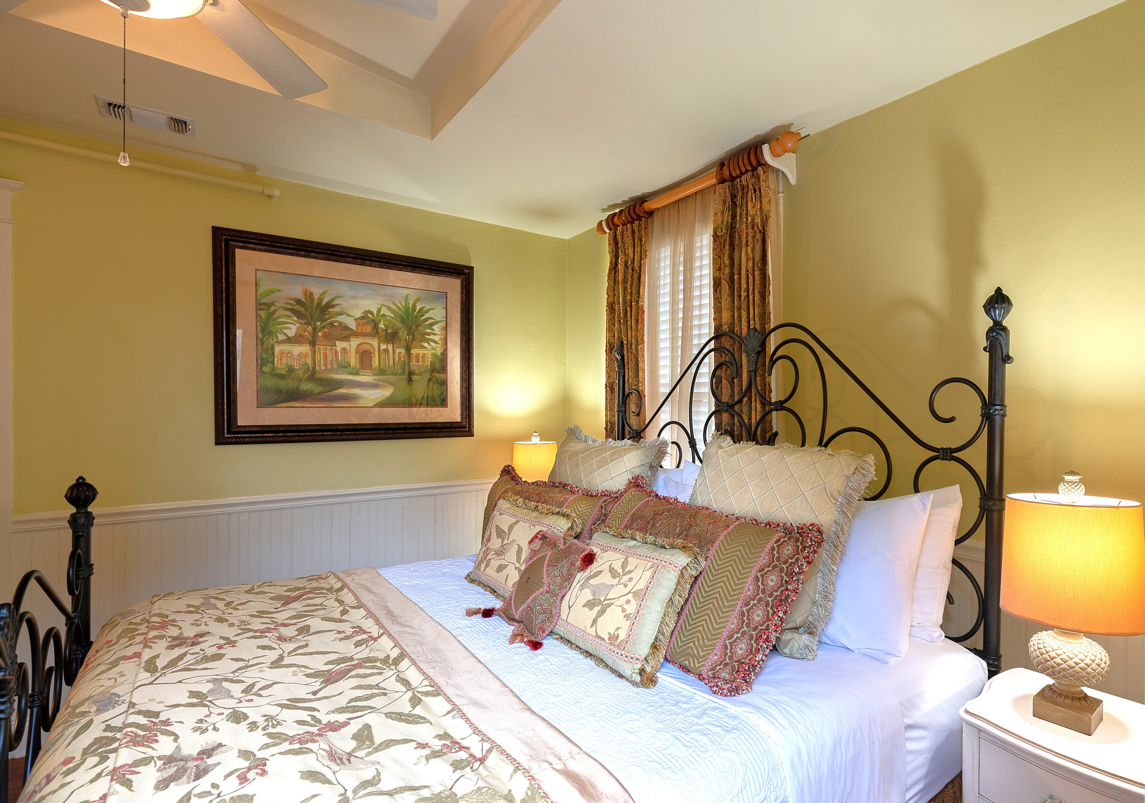 A traditionally styled bedroom with a black iron bed frame, a painting hanging above the bed, and a lamp on a nightstand.