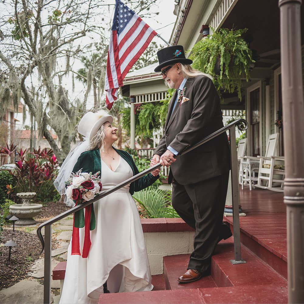 A joyful couple in wedding attire ascends outdoor steps, framed by an American flag and lush greenery.