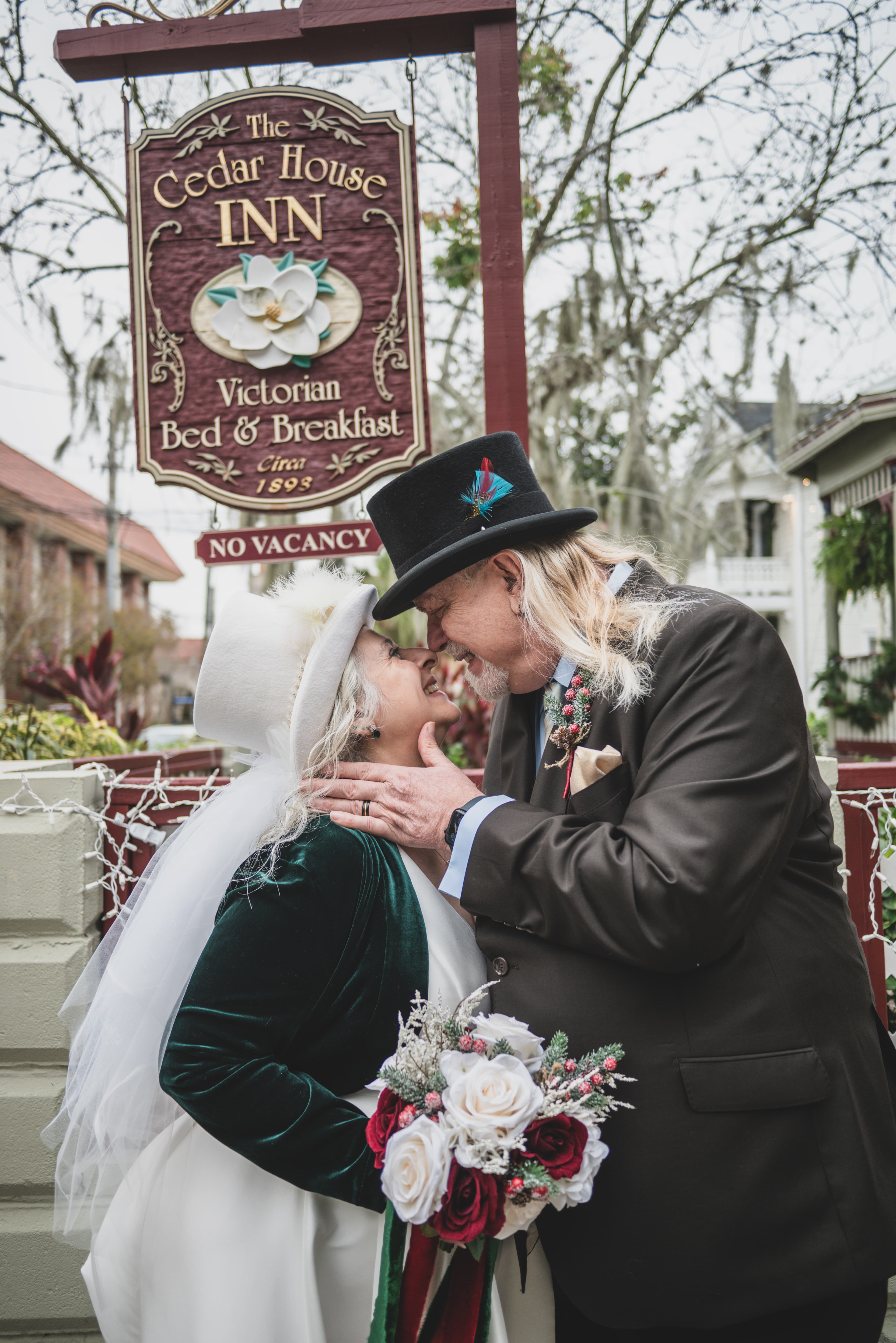 A couple in elegant attire shares a joyful moment in front of the Cedar House Inn sign.