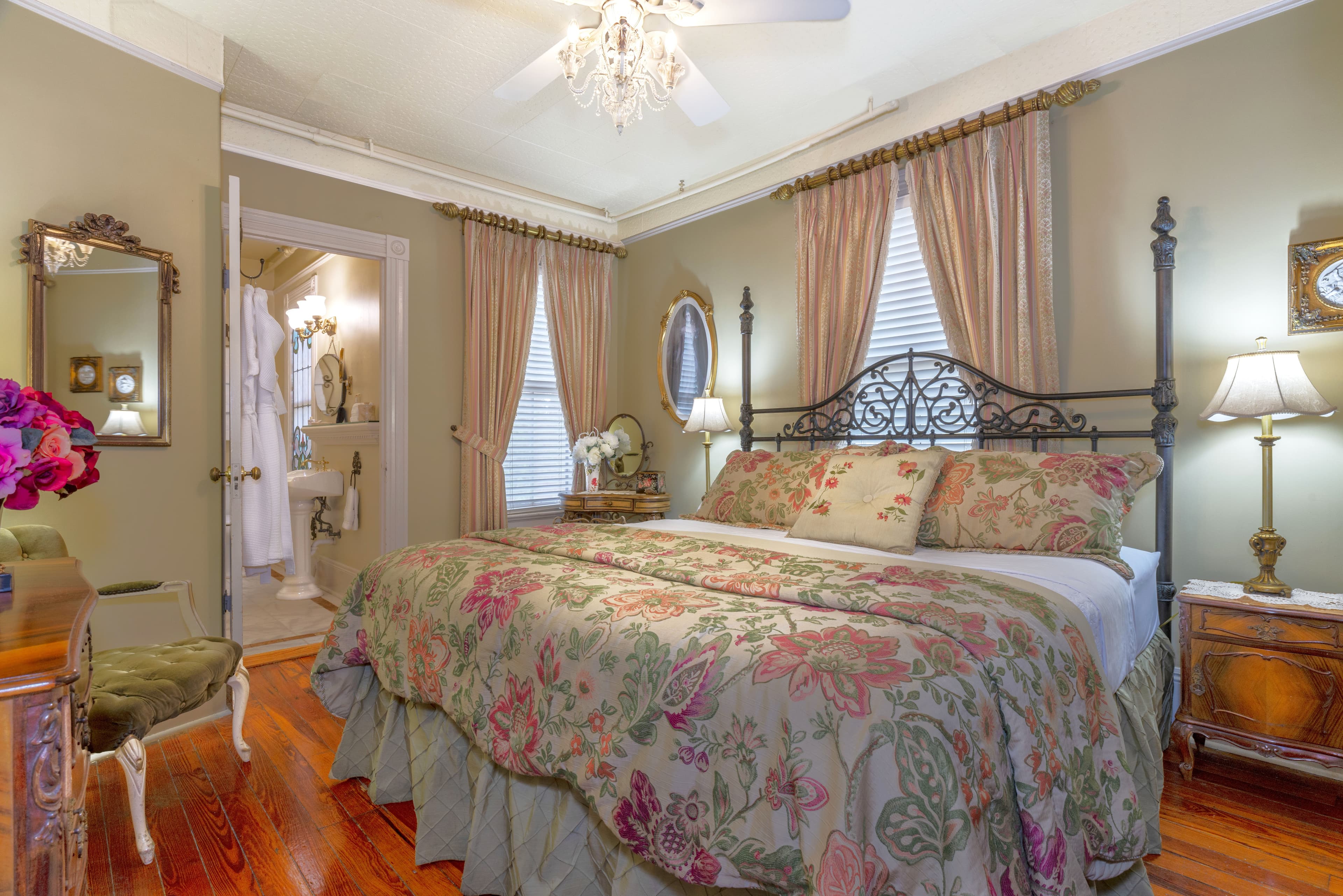 A cozy bedroom featuring a floral-patterned bedspread, elegant drapes, and a view of an adjoining bathroom.