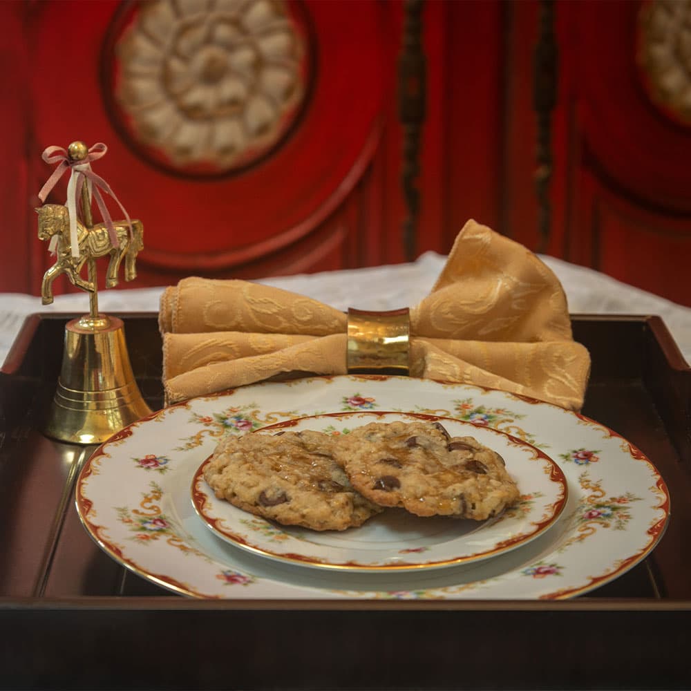 A plate of cookies sits on an ornate tray beside a golden bell and a neatly tied golden napkin.