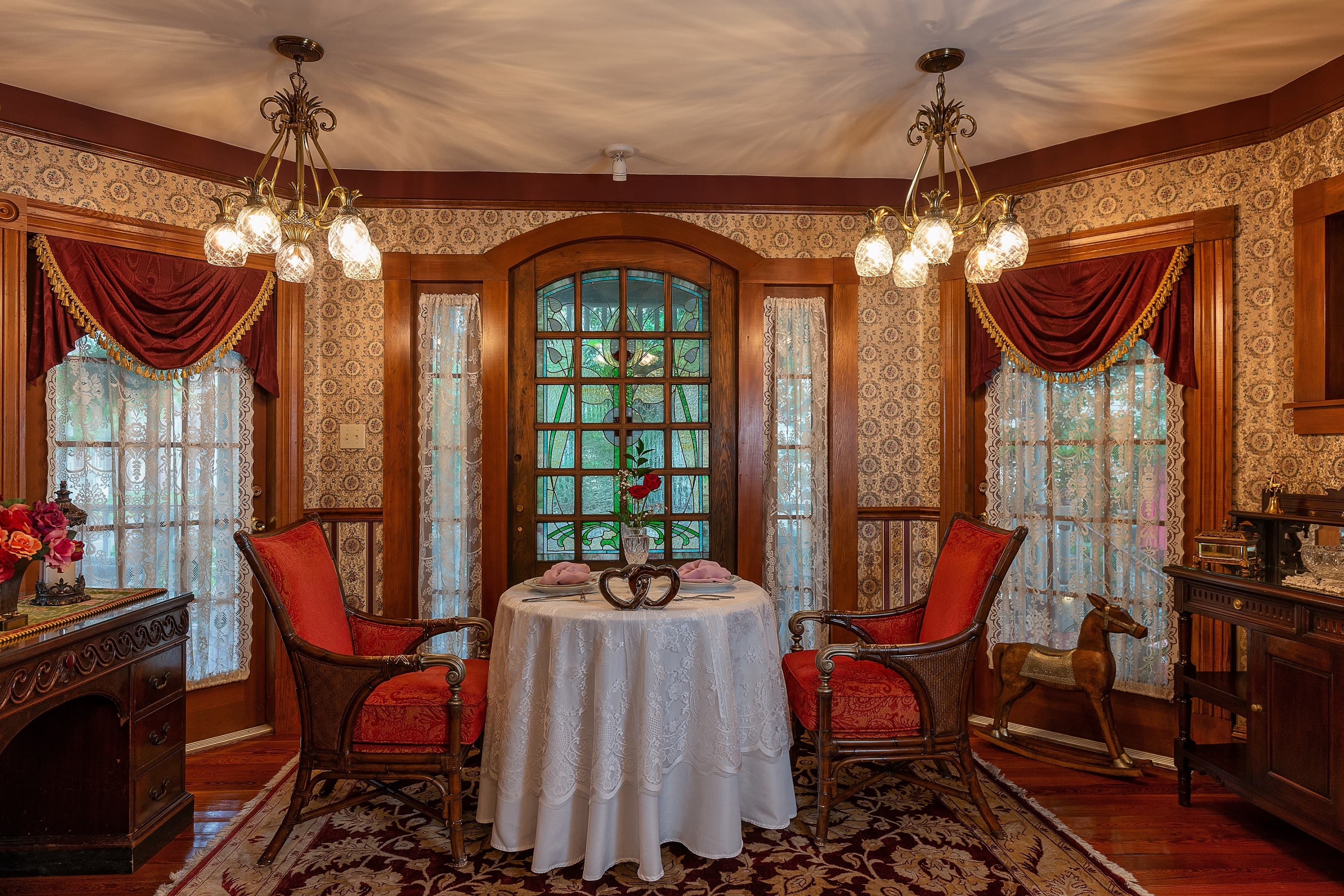 A cozy dining nook featuring a round table set with pink napkins, two red chairs, and decorative vintage accents against a floral wallpaper backdrop.
