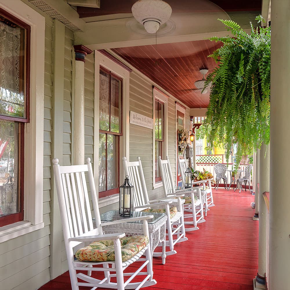 A cozy porch with rocking chairs and greenery, featuring red flooring and decorative elements.