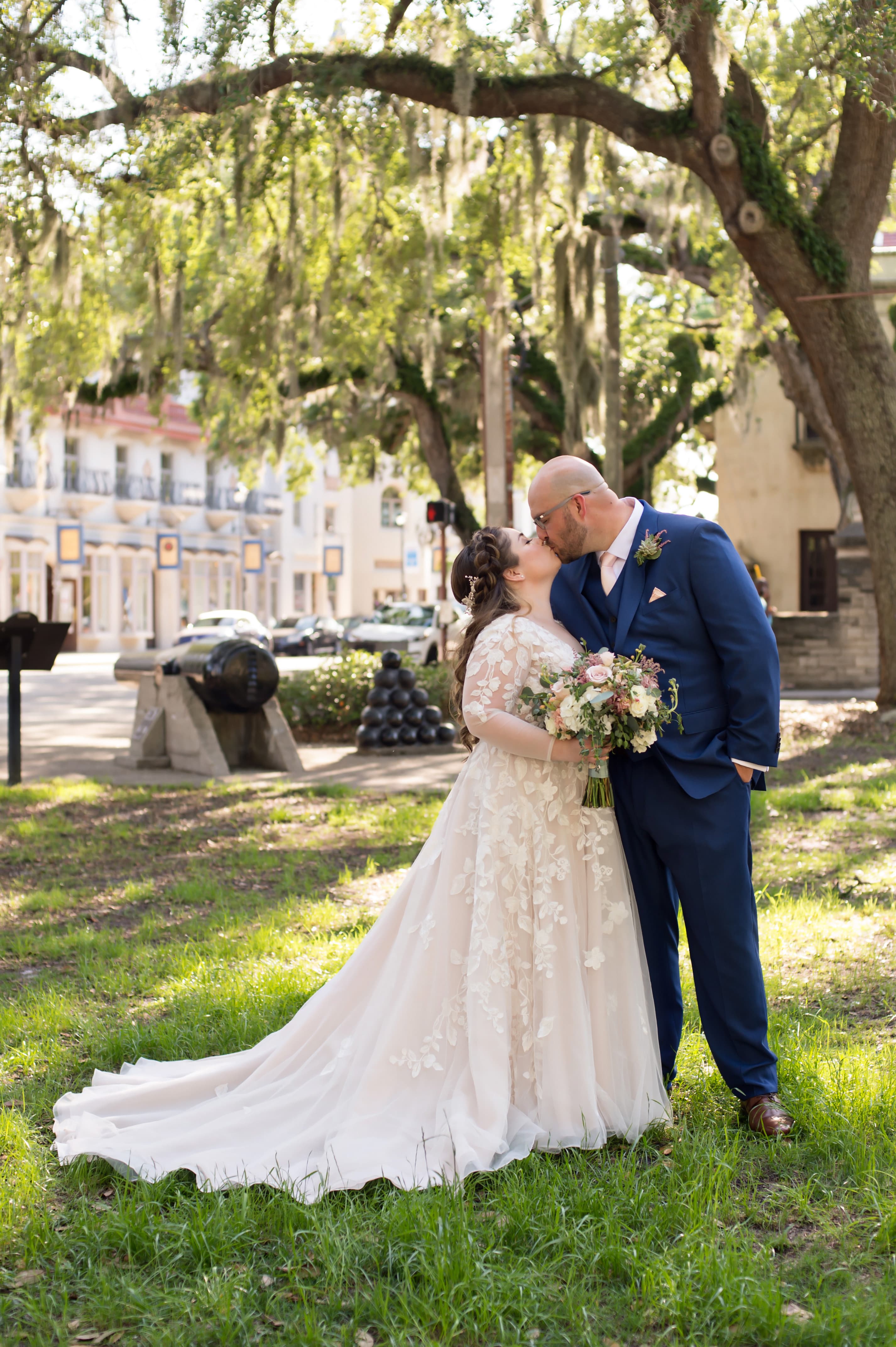 A bride and groom kiss in a lush park, surrounded by greenery and historic architecture.
