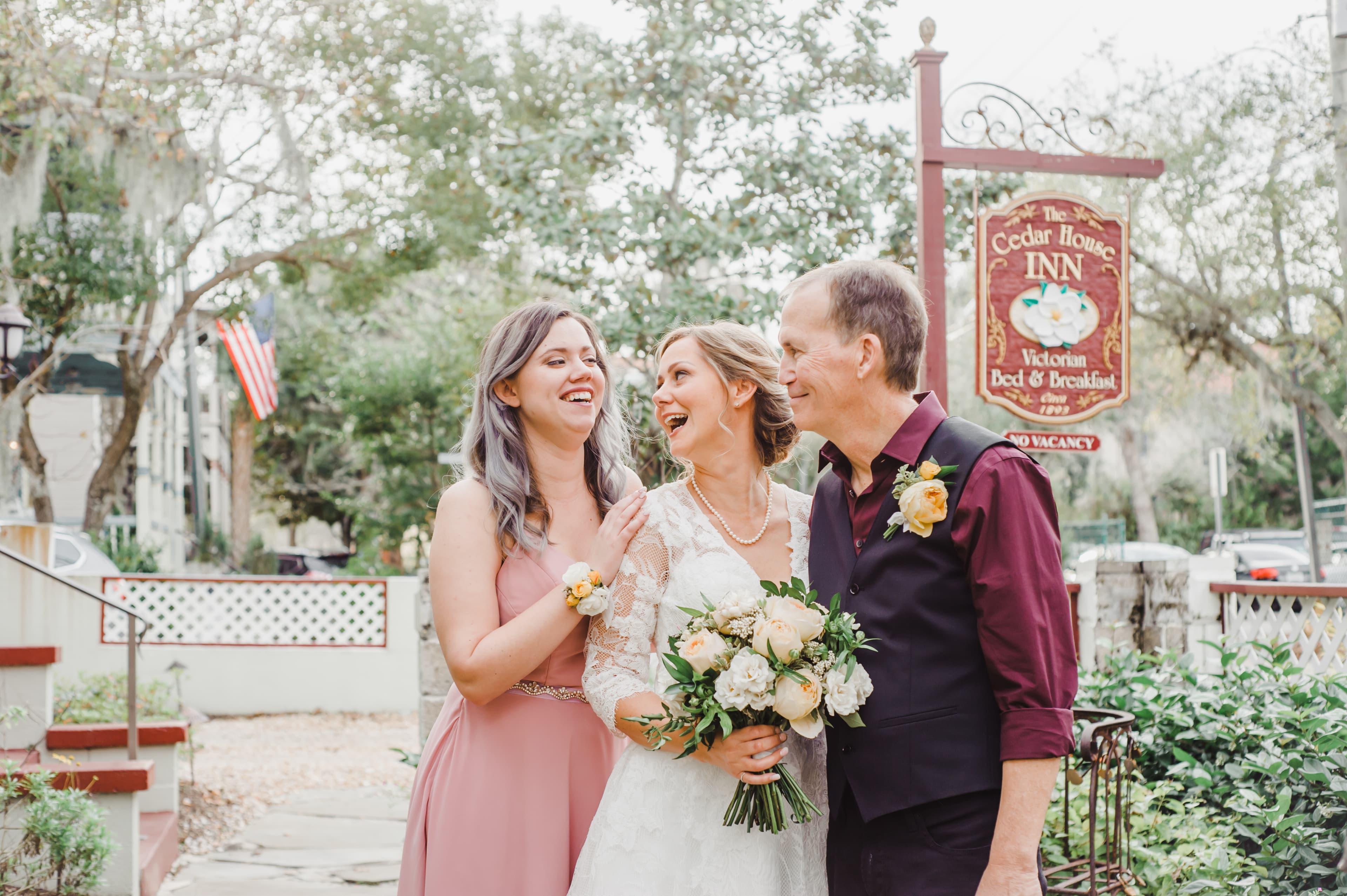 A bride smiles joyfully with two guests in a garden setting near a sign for The Cedar House Inn.