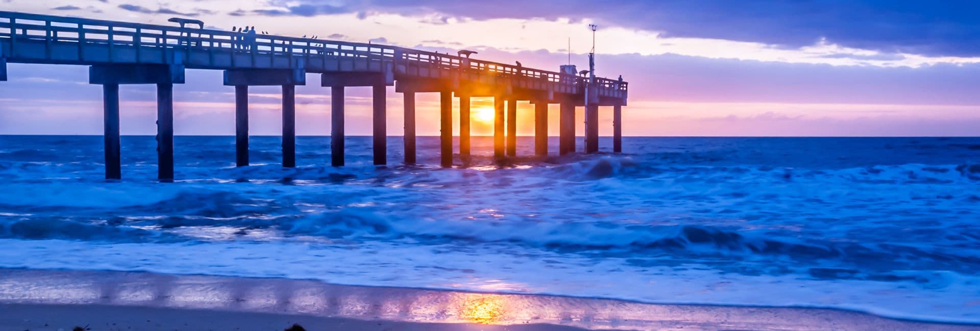 A sunset glows beneath a pier, casting reflections on the ocean waves.