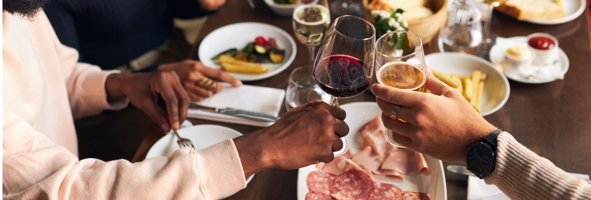 Two hands clink glasses of red wine and beer over a table set with various dishes.