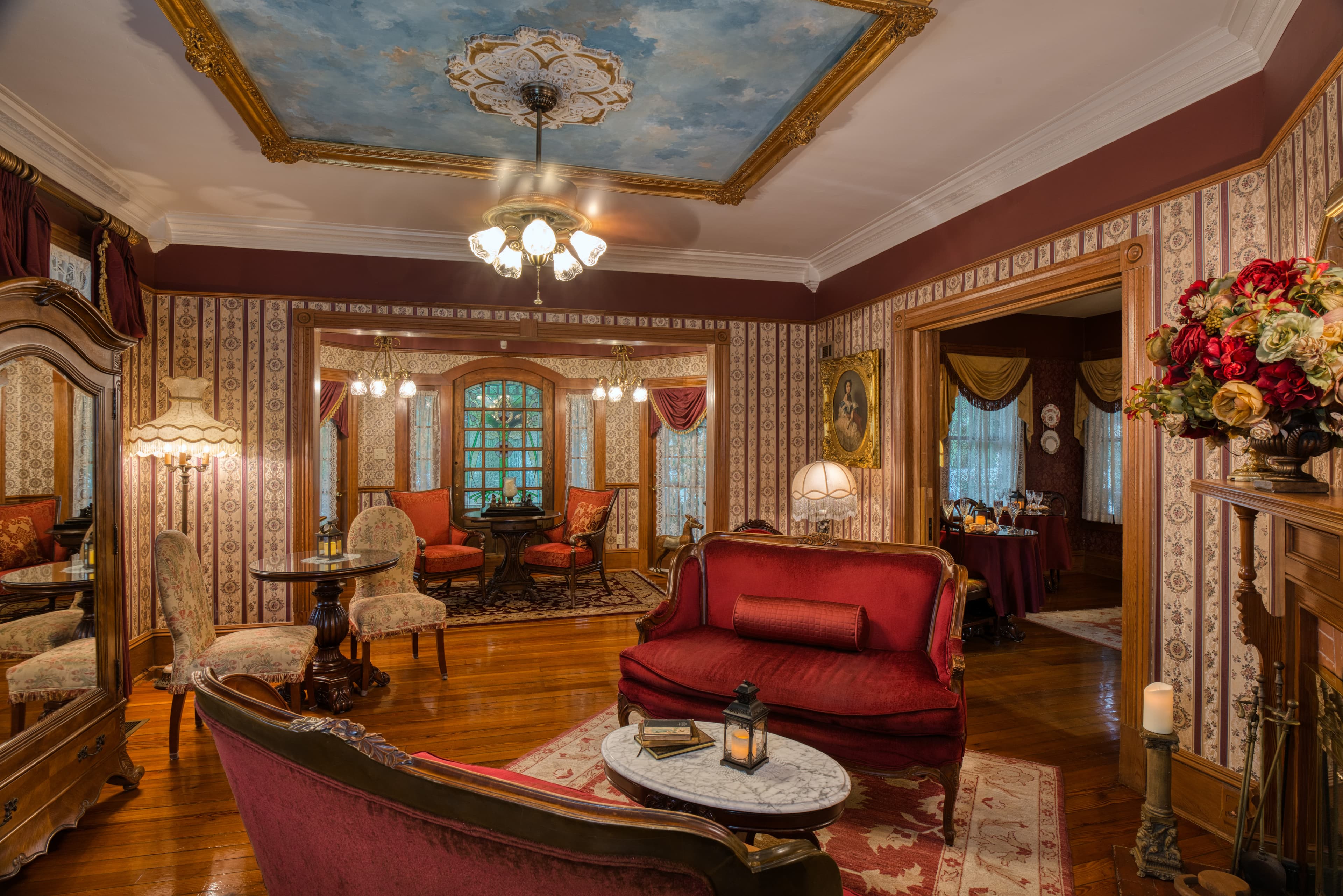 A plush, vintage sitting room featuring ornate wallpaper, a decorative ceiling, and elegant furnishings in rich colors.