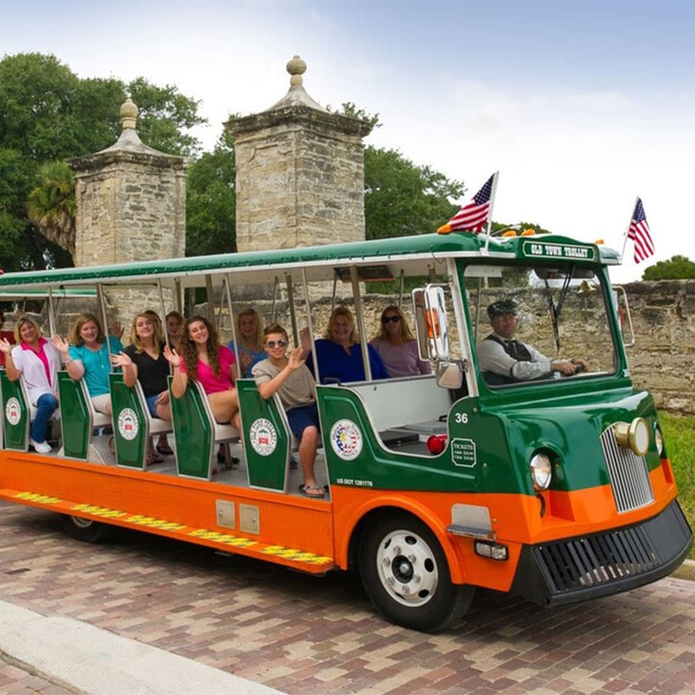 A green and orange tourist trolley with passengers waves as it stands in front of historic stone towers.