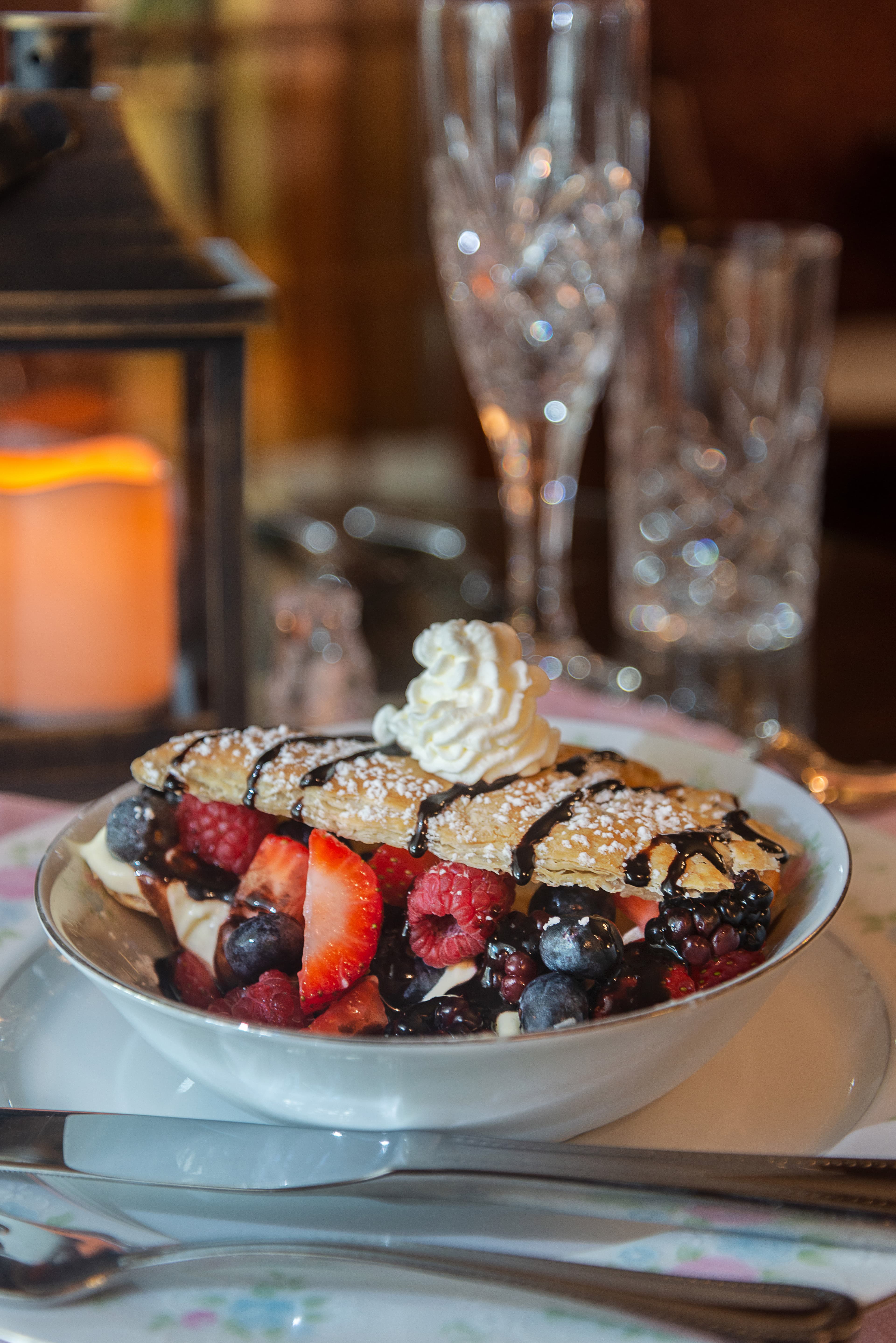 A dessert bowl filled with mixed berries, chocolate drizzle, and whipped cream, beside elegant glassware and a candle.