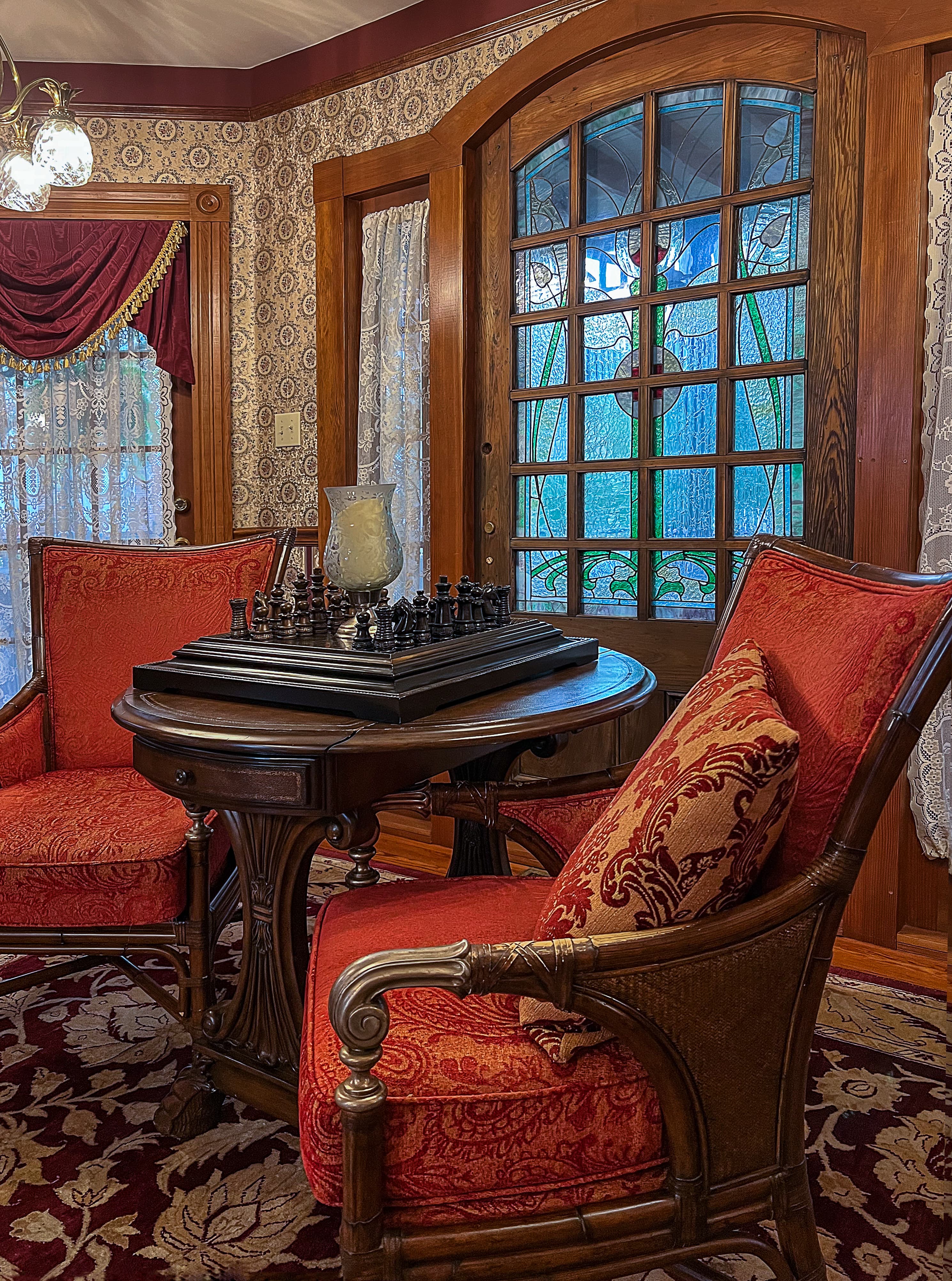 A cozy parlor with two red upholstered chairs, a wooden chess table, and a stained glass door.