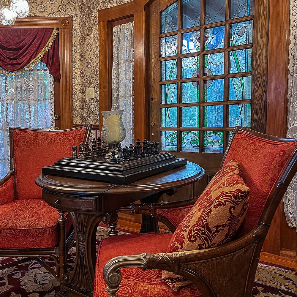 A cozy sitting area with red chairs, a chess set on a dark wooden table, and a stained glass door.
