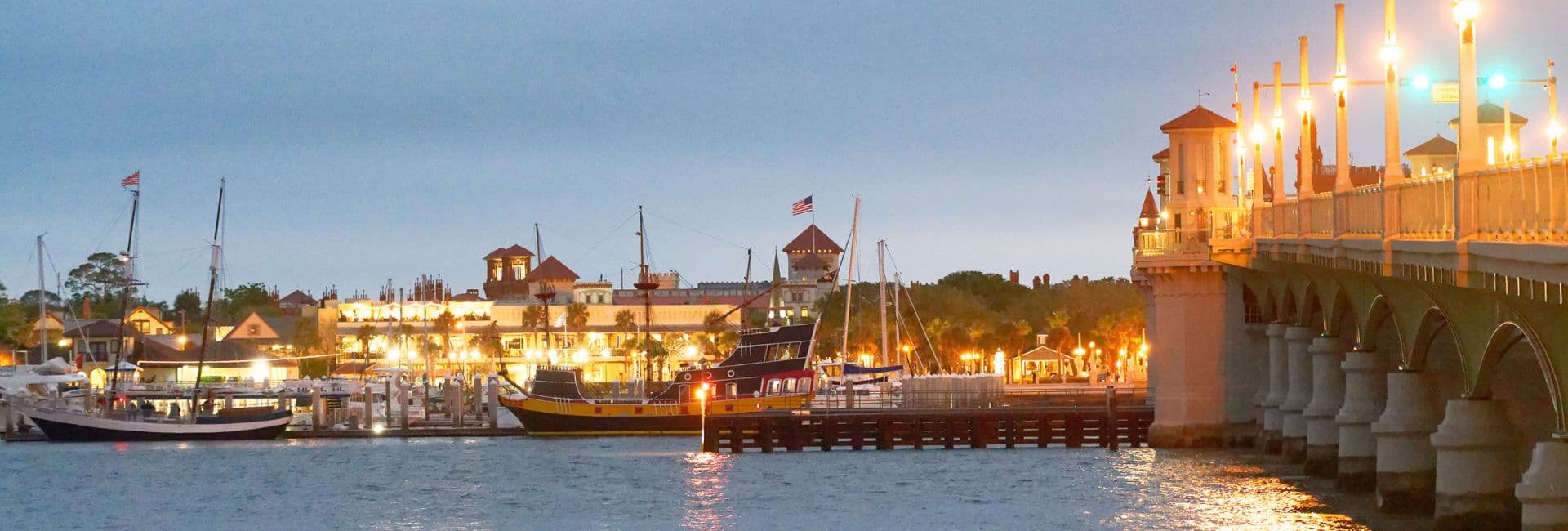 A view of a harbor at dusk featuring illuminated buildings, boats, and a bridge.