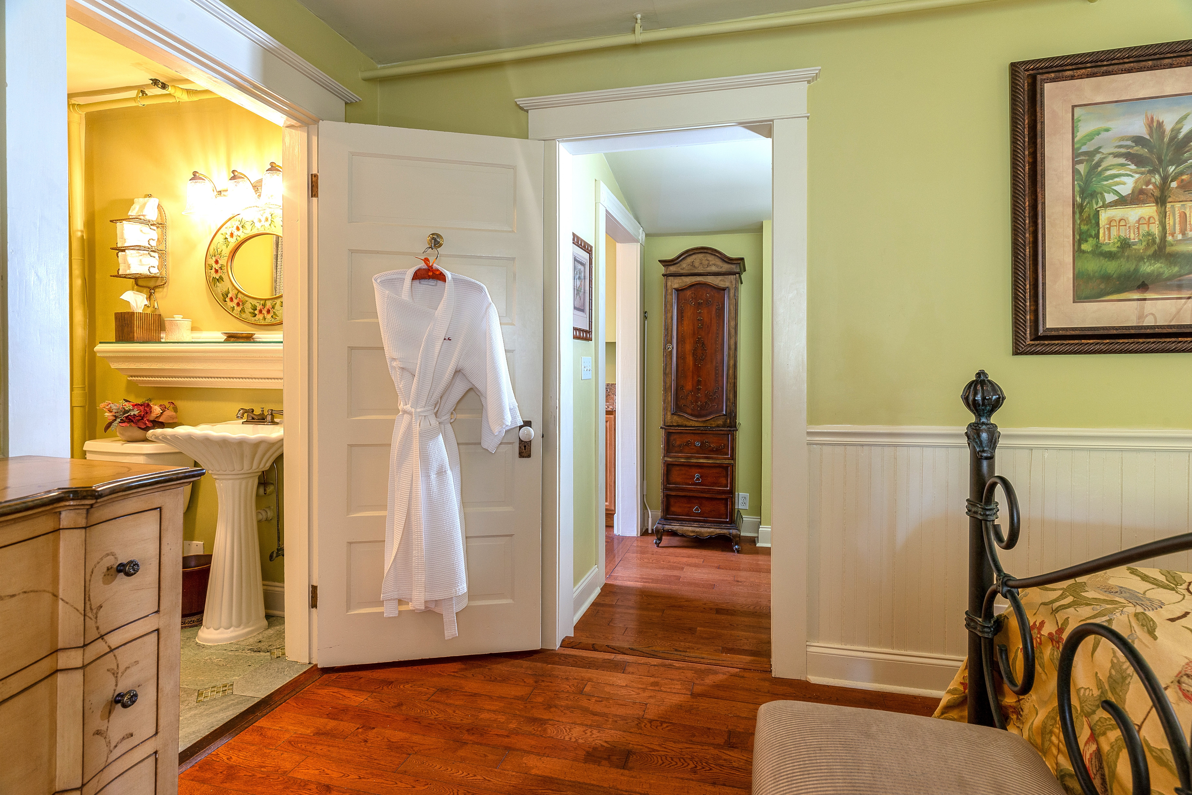 A hotel room with a dark wood bed frame, a bathroom with a sink and a mirror, and a hallway leading to another room. A white bathrobe hangs on the bathroom door.