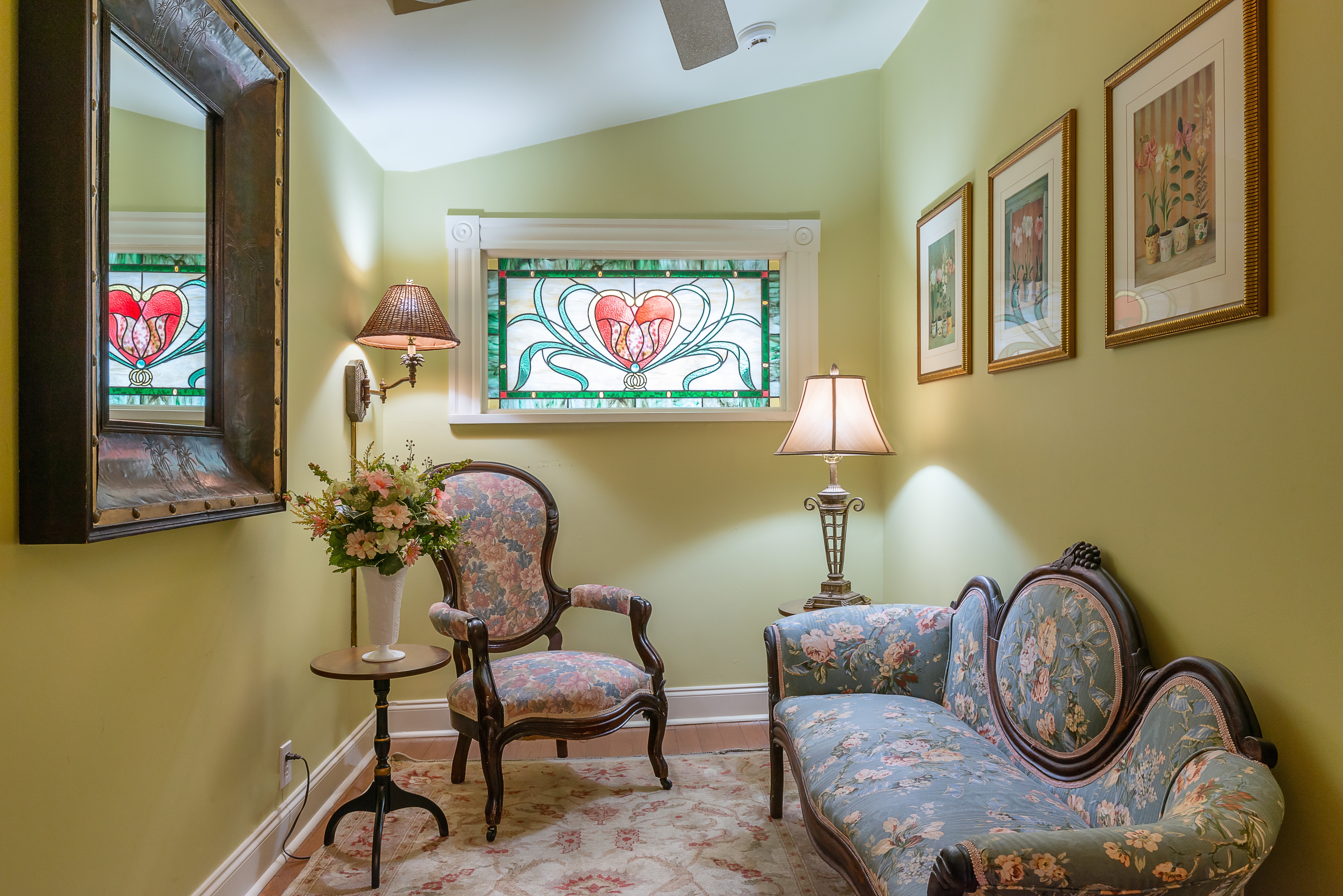 A traditionally decorated sitting room featuring a patterned sofa and chair, a small table with a flower arrangement, and a stained glass window with a red heart design.