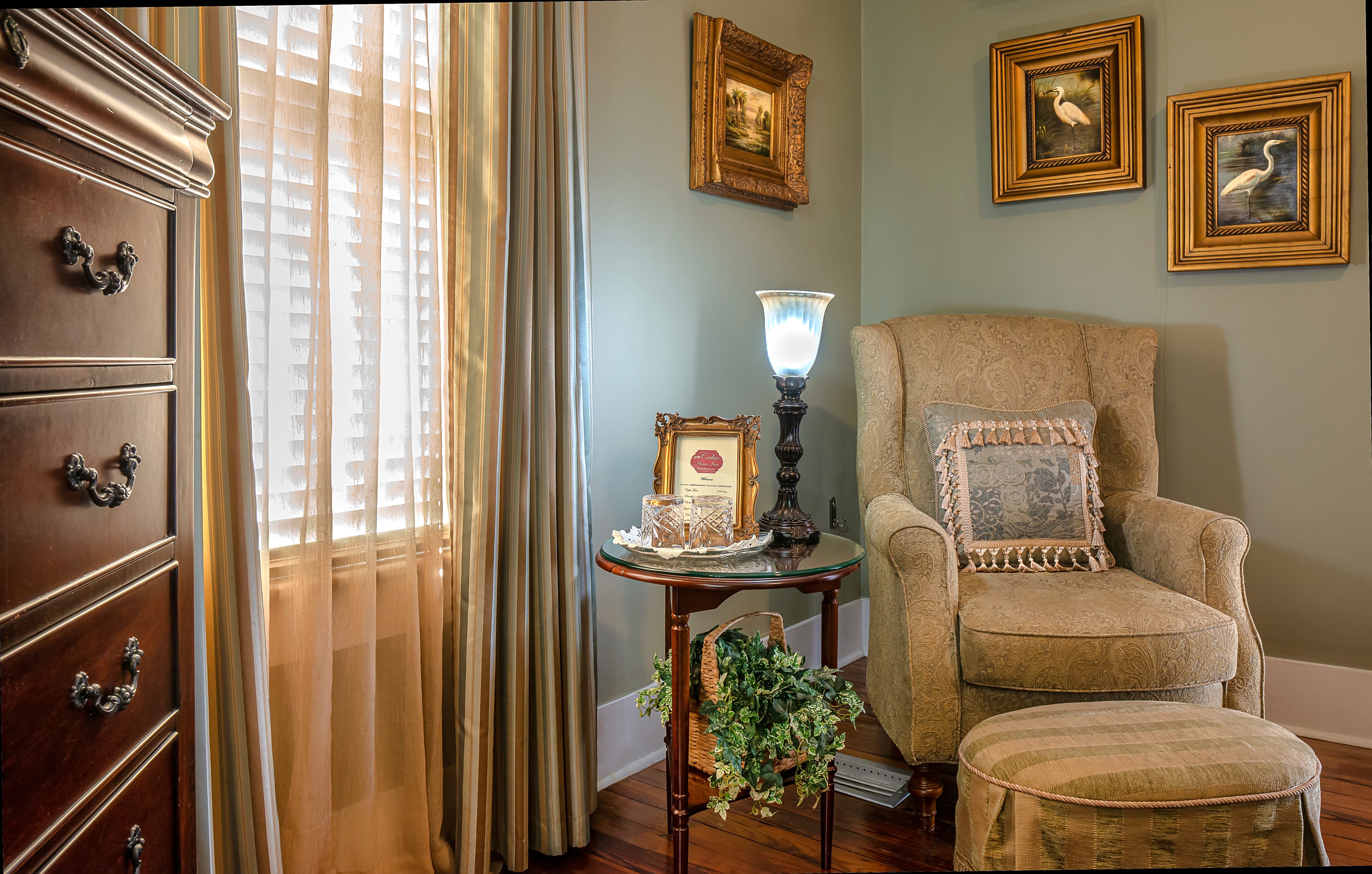 A comfortable armchair and ottoman in a sitting area with a small table and a lamp. Three framed pictures hang on the wall, and a large window with a light-colored curtain is visible on the left.