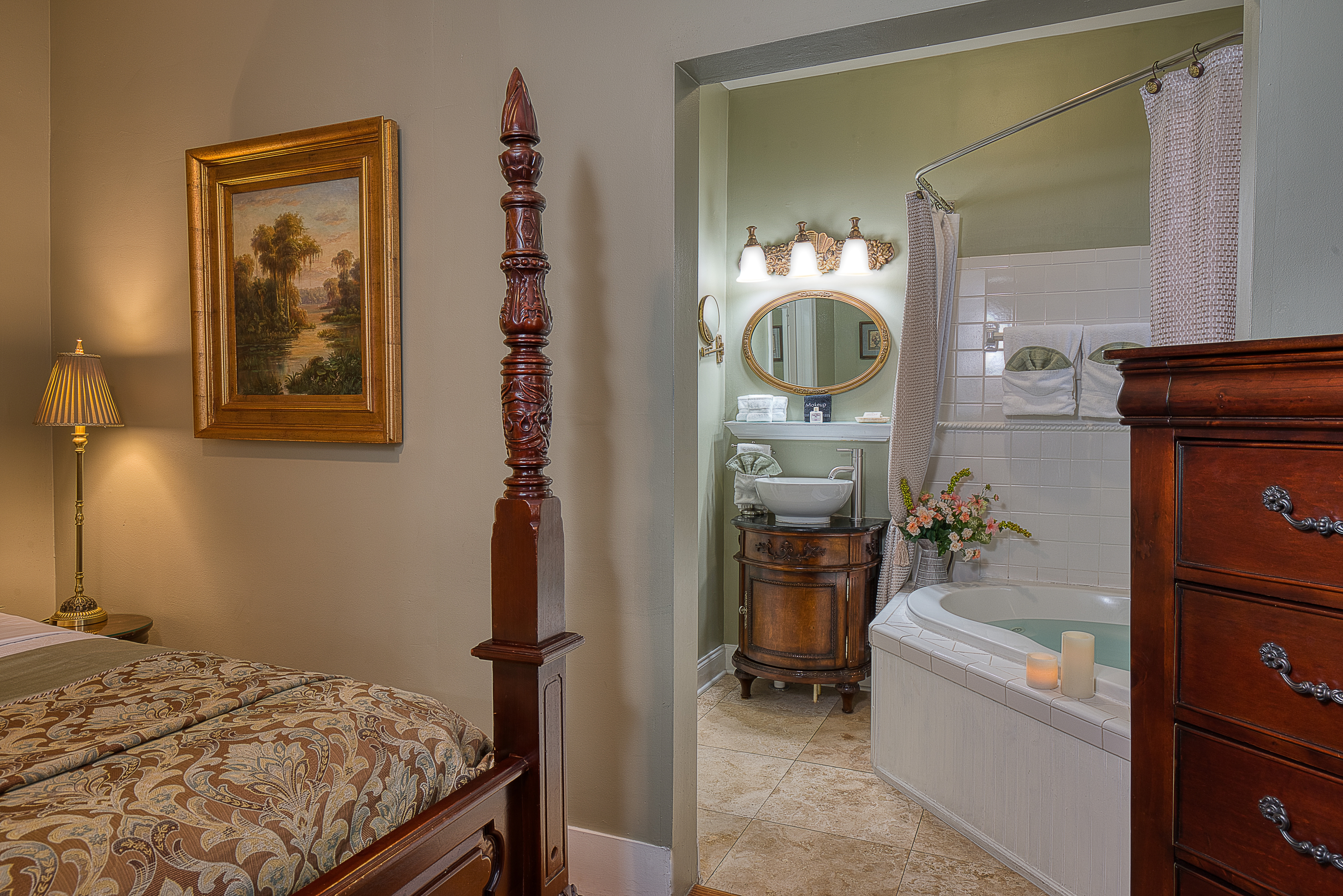 A lovely room with a four-poster bed, a dark wood dresser, and a bathroom with a jacuzzi tub and a sink. A framed picture hangs on the wall above the bed.