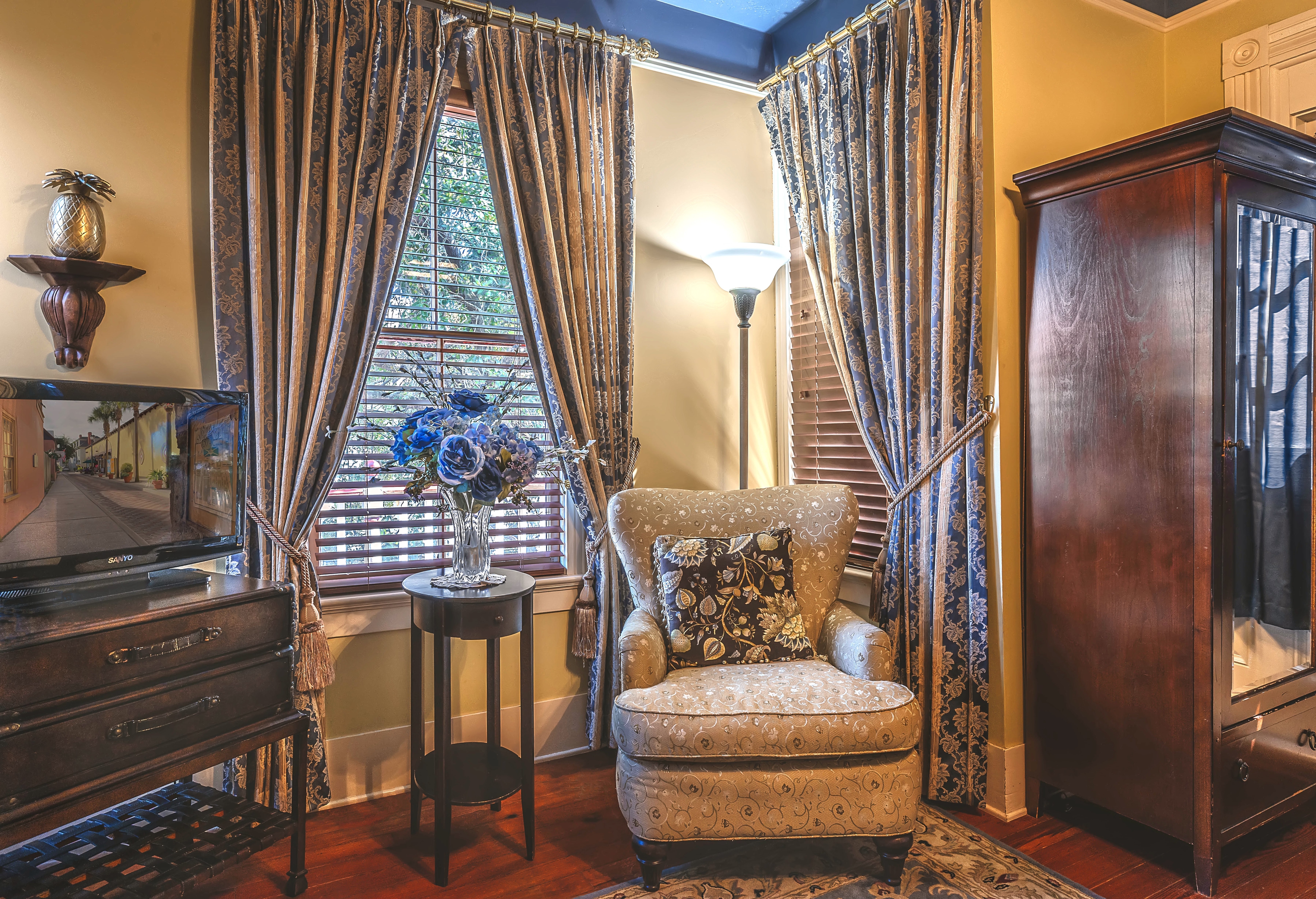 A sitting area in a bedroom with a patterned armchair, a small table with a lamp, and a large window.