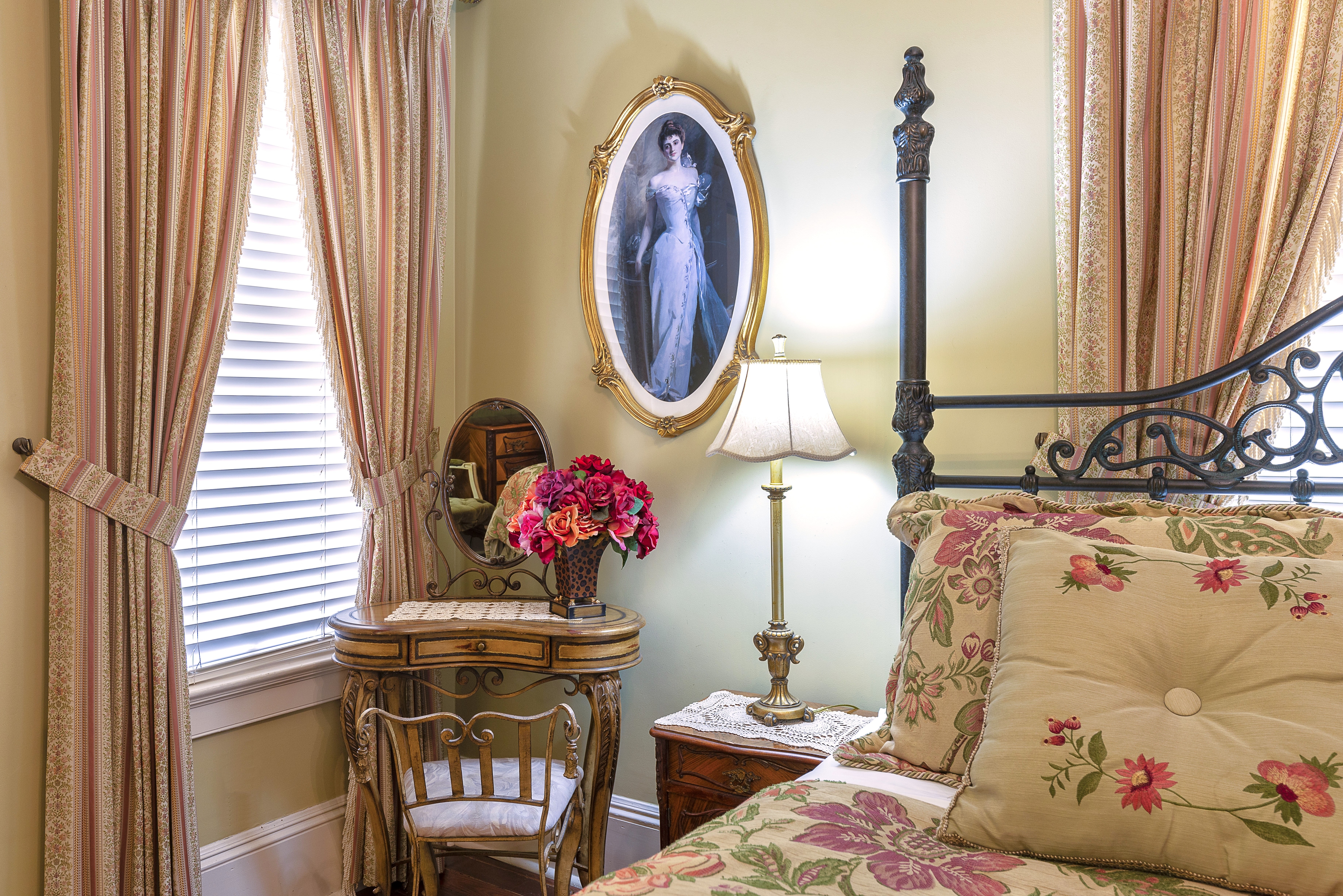 An elegant bedroom featuring a bed with a patterned comforter and pillows, a vanity with a mirror and a chair, a sitting area with a lamp, and a sunny window.