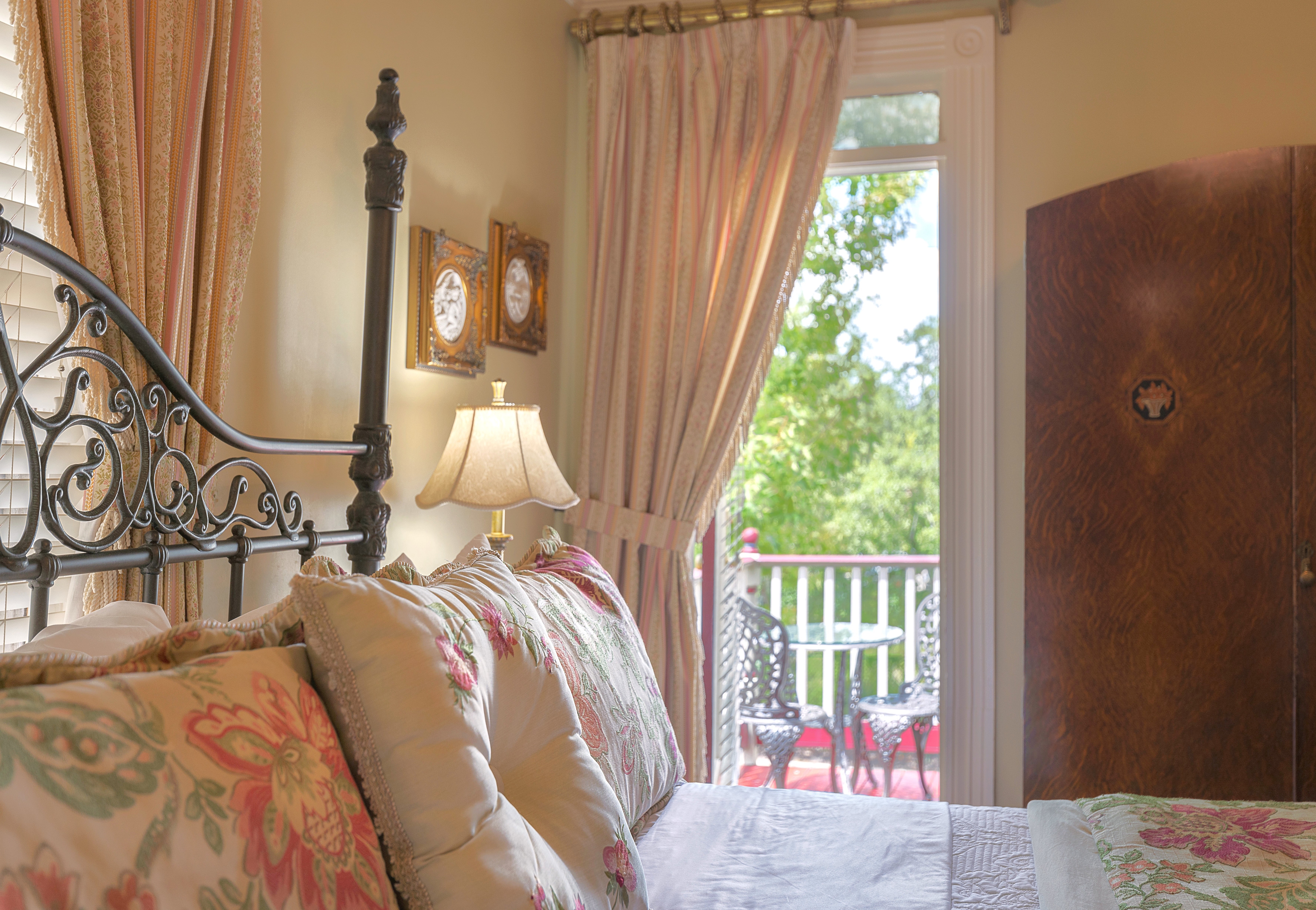 Romantic room featuring a bed with a patterned comforter and pillows, a dresser, a sitting area with a chair and a lamp, and a balcony with a view of trees.