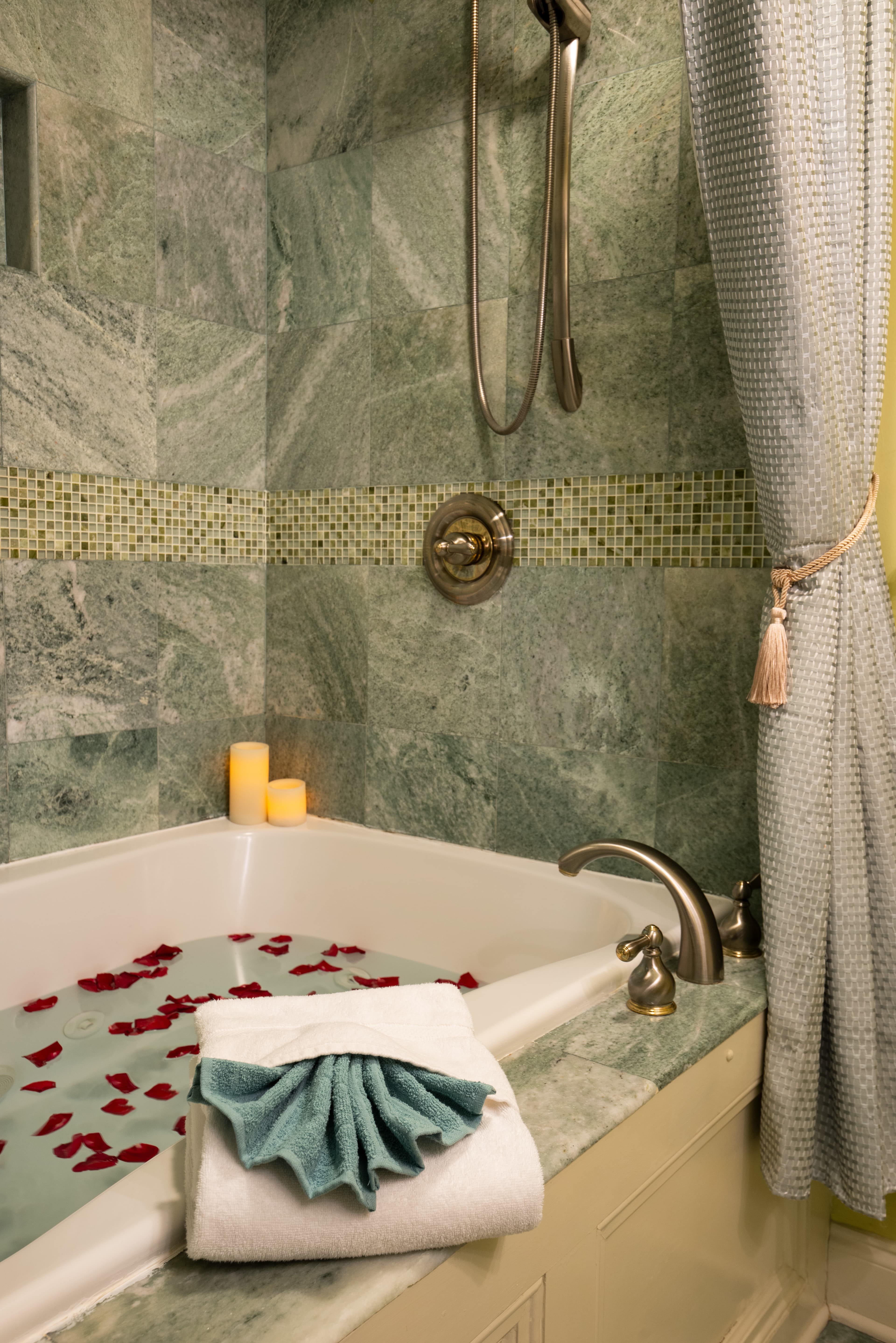 A close-up view of a bathtub with a shower head and a shower curtain. A folded towel is placed on the edge of the tub, and rose petals are scattered in the water.