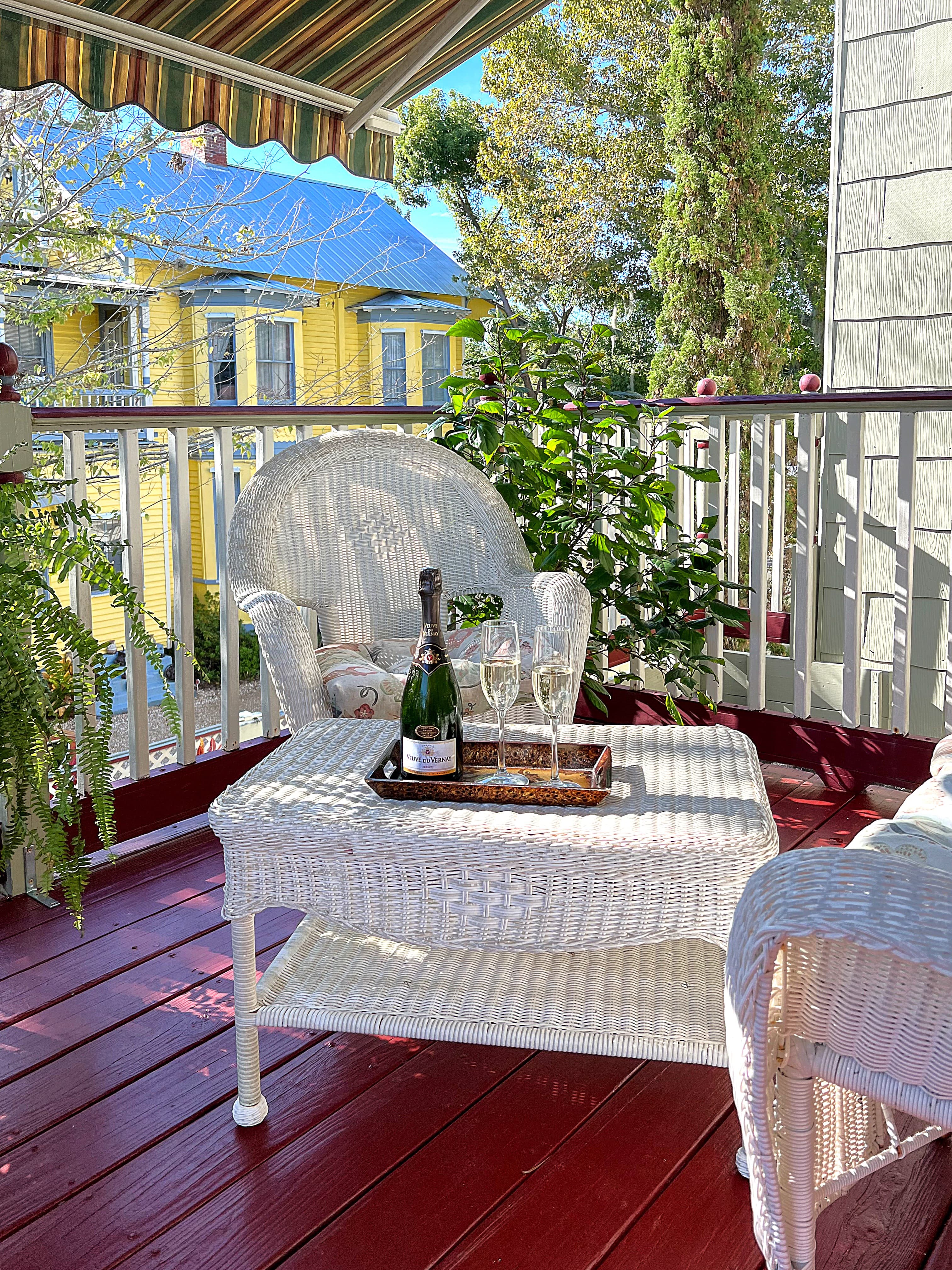 A white wicker chair and ottoman on a porch with a bottle of champagne and two glasses on a tray. The porch overlooks a colorful building and trees.