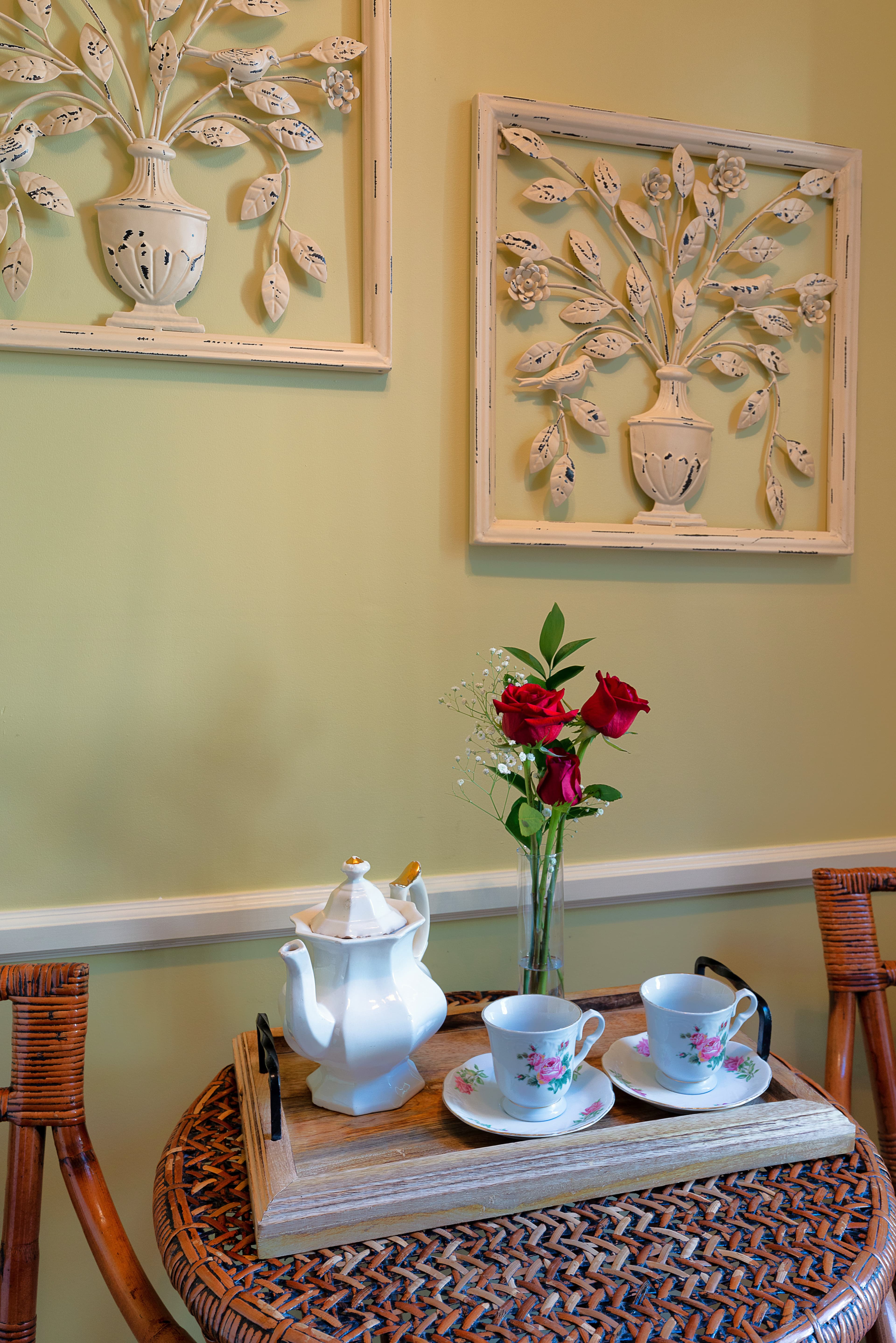 A round wicker table with a tea set and a vase of red roses, with two decorative wall art pieces hanging above.