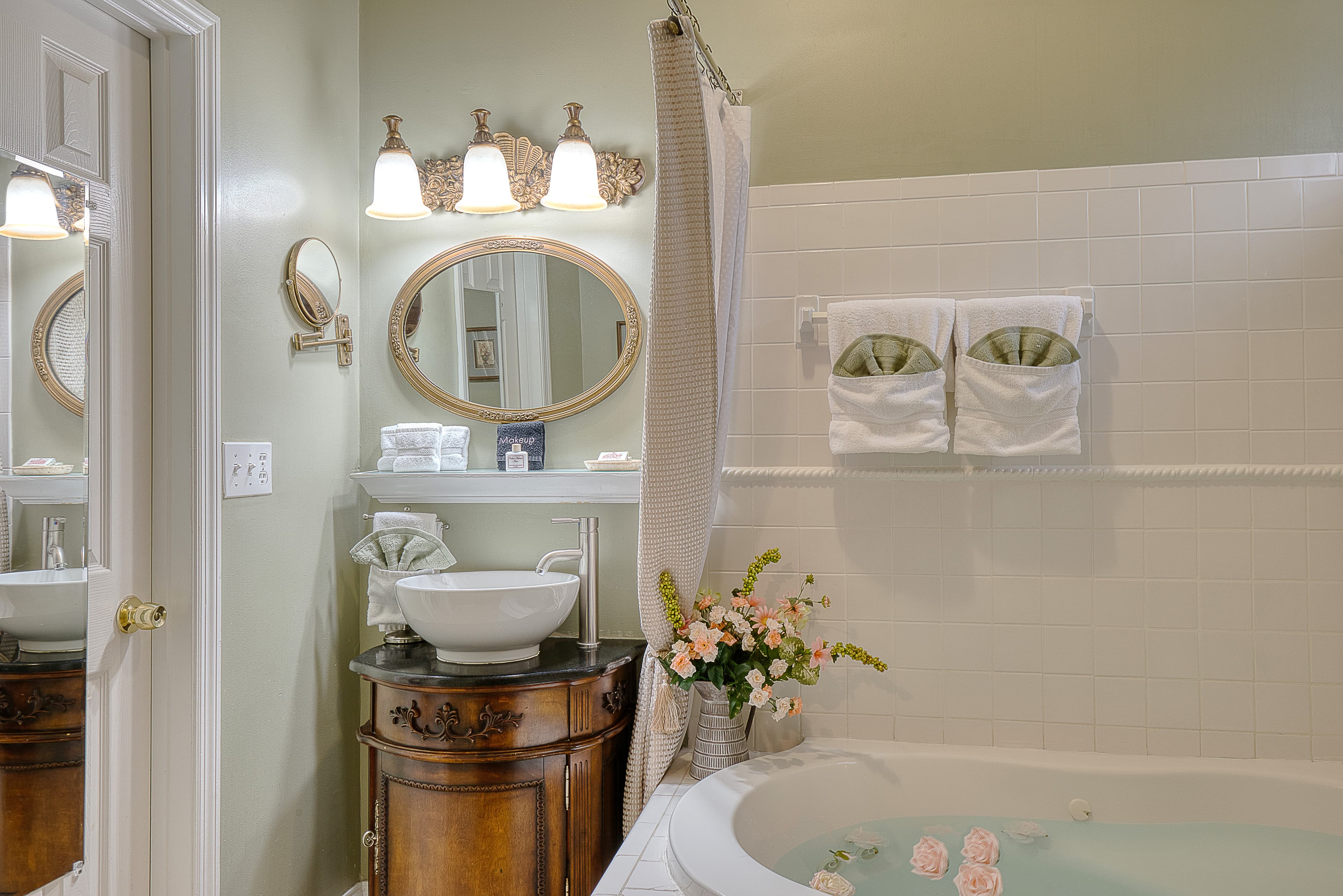 Interior view of a bathroom featuring a jacuzzi tub with rose petals, a sink on a dark wood vanity with a decorative mirror above it, and a light fixture.