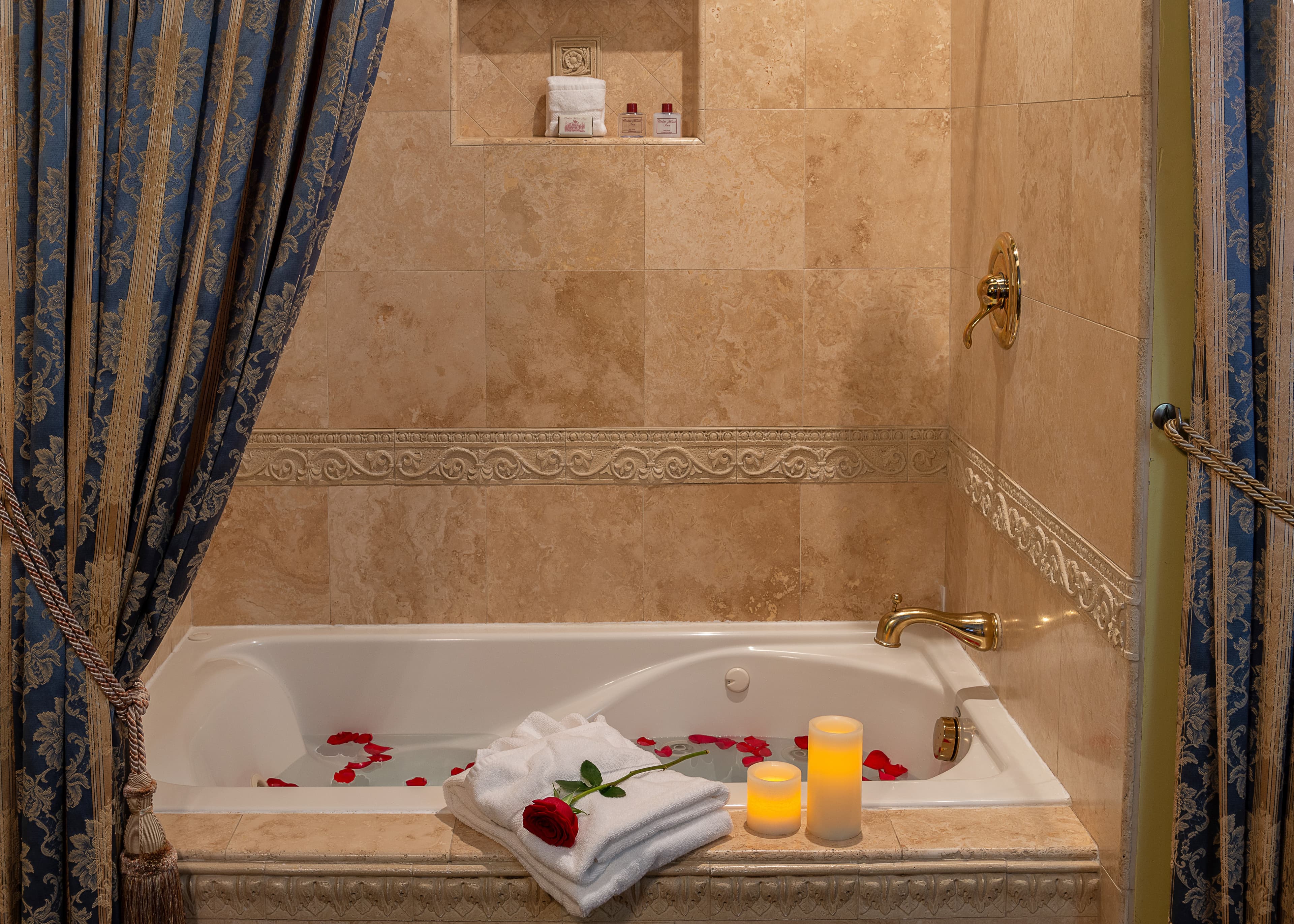 Close-up view of a bathtub with rose petals scattered in the water, a folded towel on the edge of the tub, and candles on a nearby shelf.