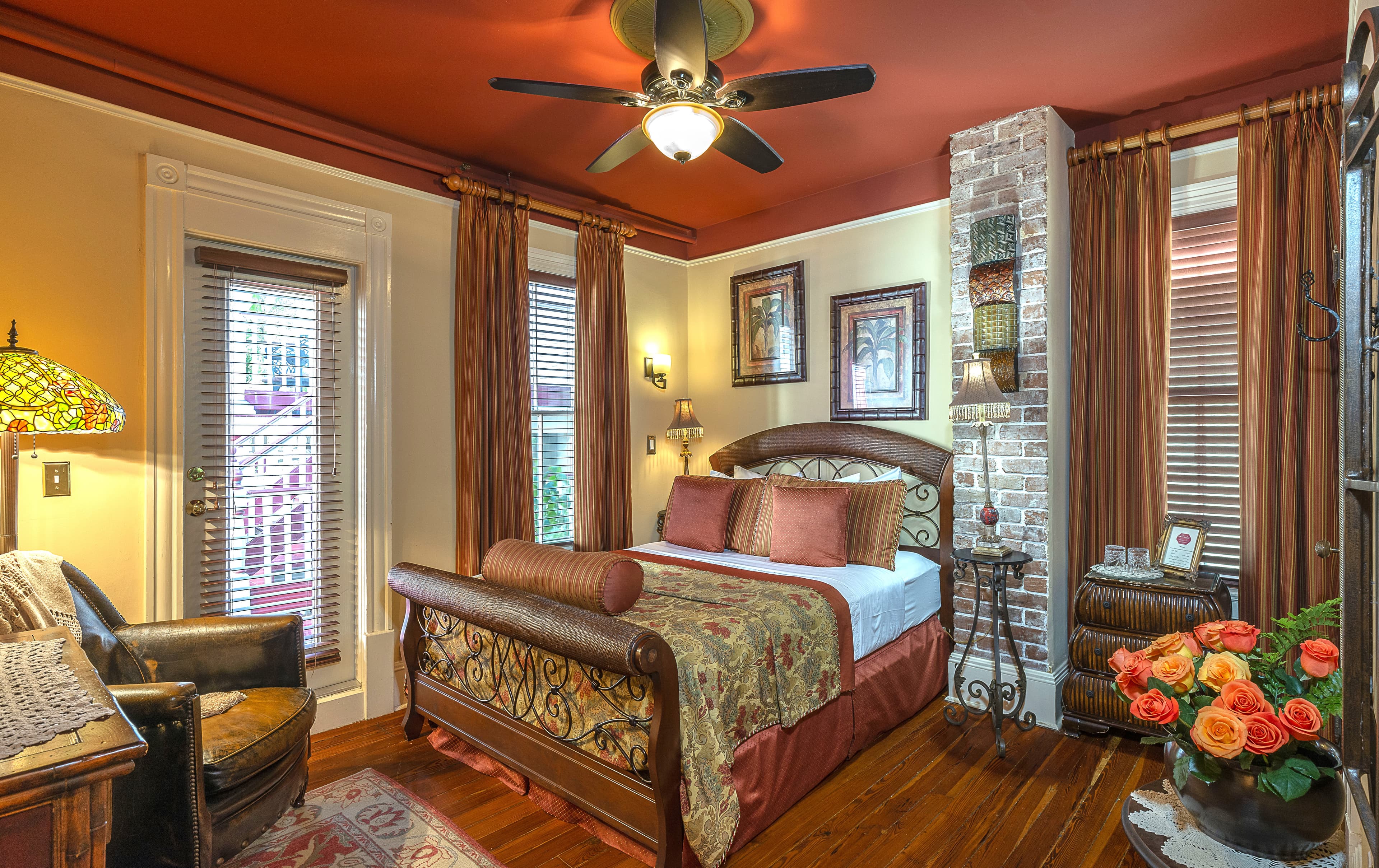 Luxurious bedroom featuring a bed with a patterned comforter and pillows, a sitting area with a chair and a lamp, and a door leading to a wrap-around porch.