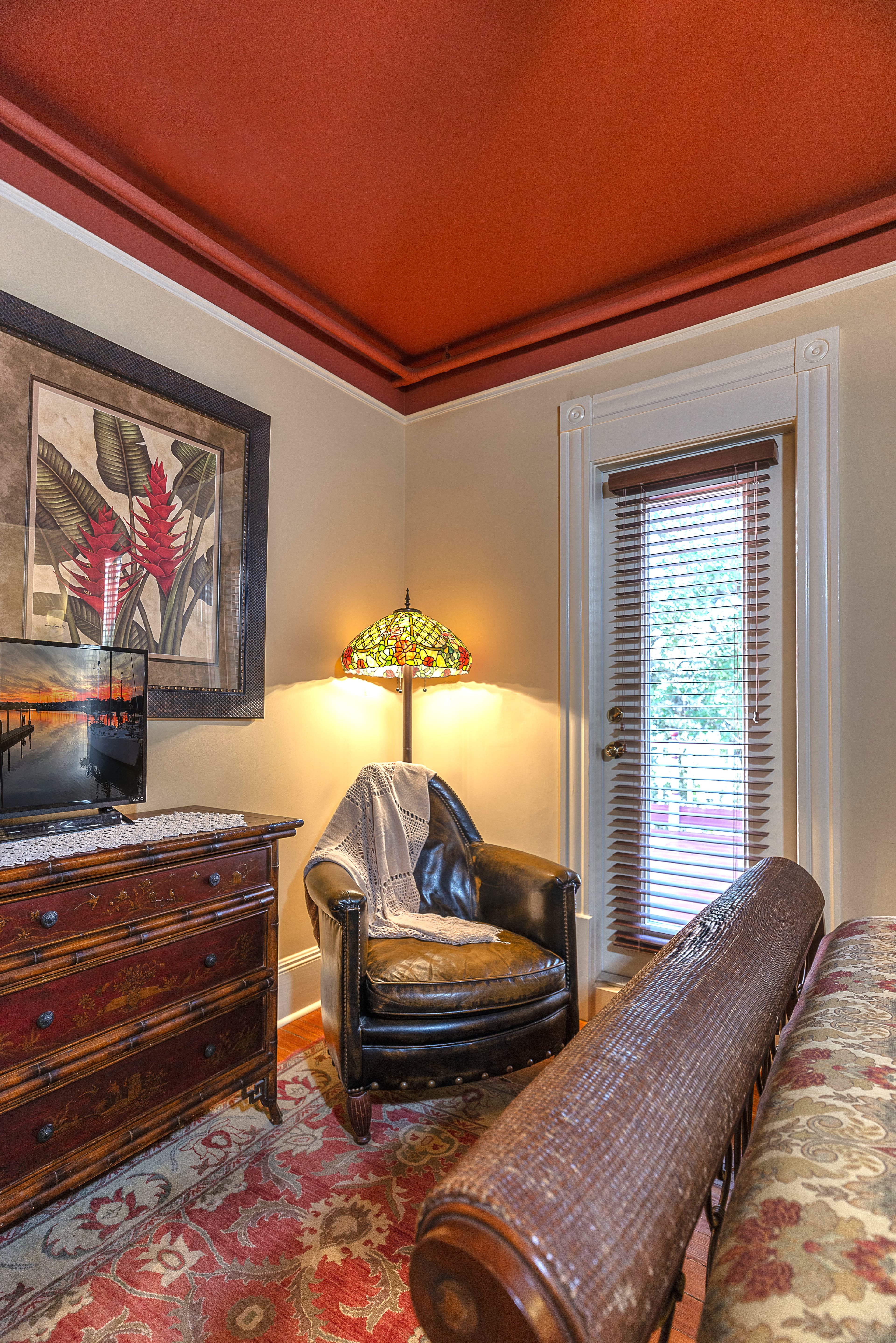 An elegant room with a bed, a dresser, a sitting area with a chair and a lamp, and a balcony with a view of trees.