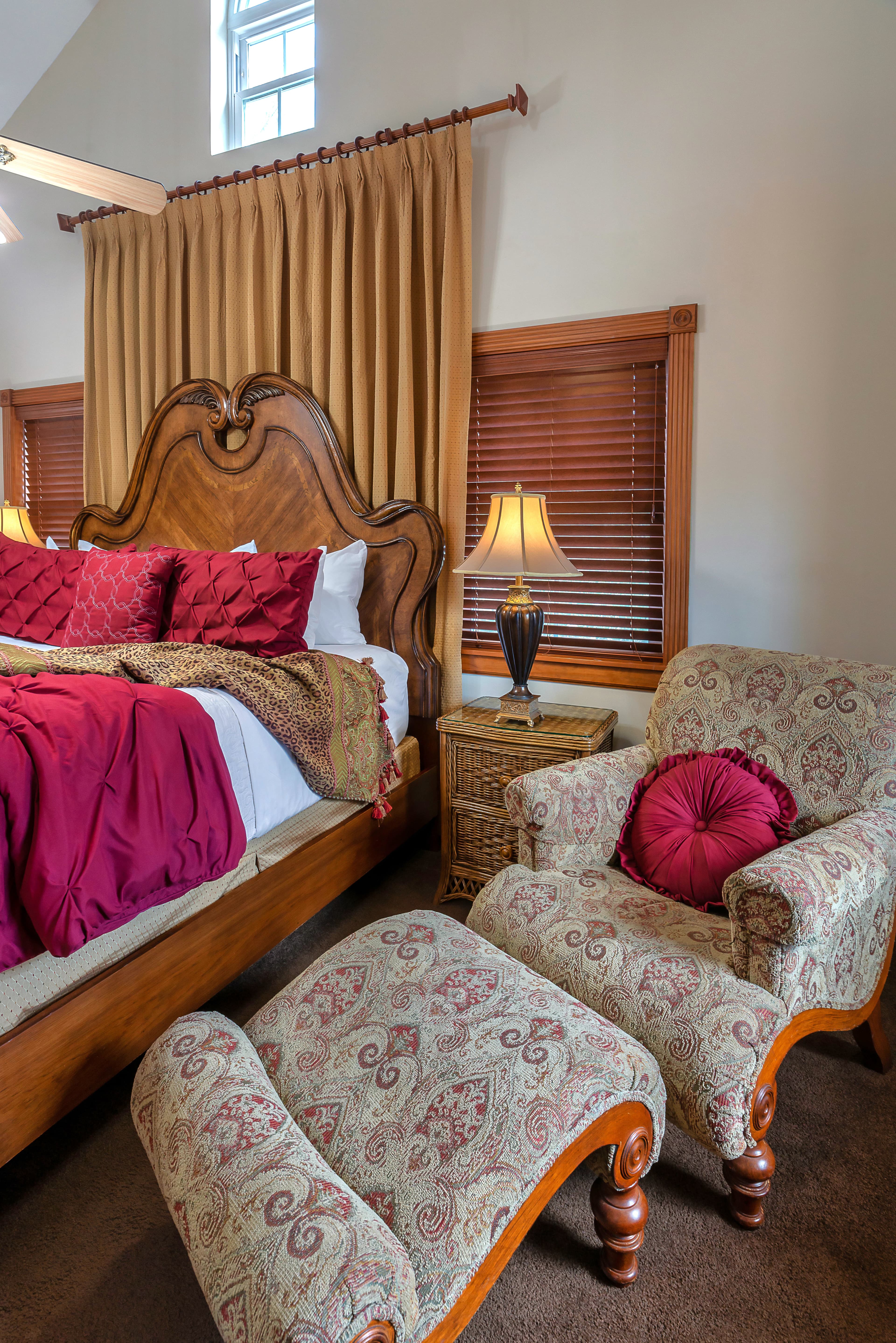 A bed with a patterned comforter and pillows, a dresser, and a sitting area with a chair, footstool, and a lamp