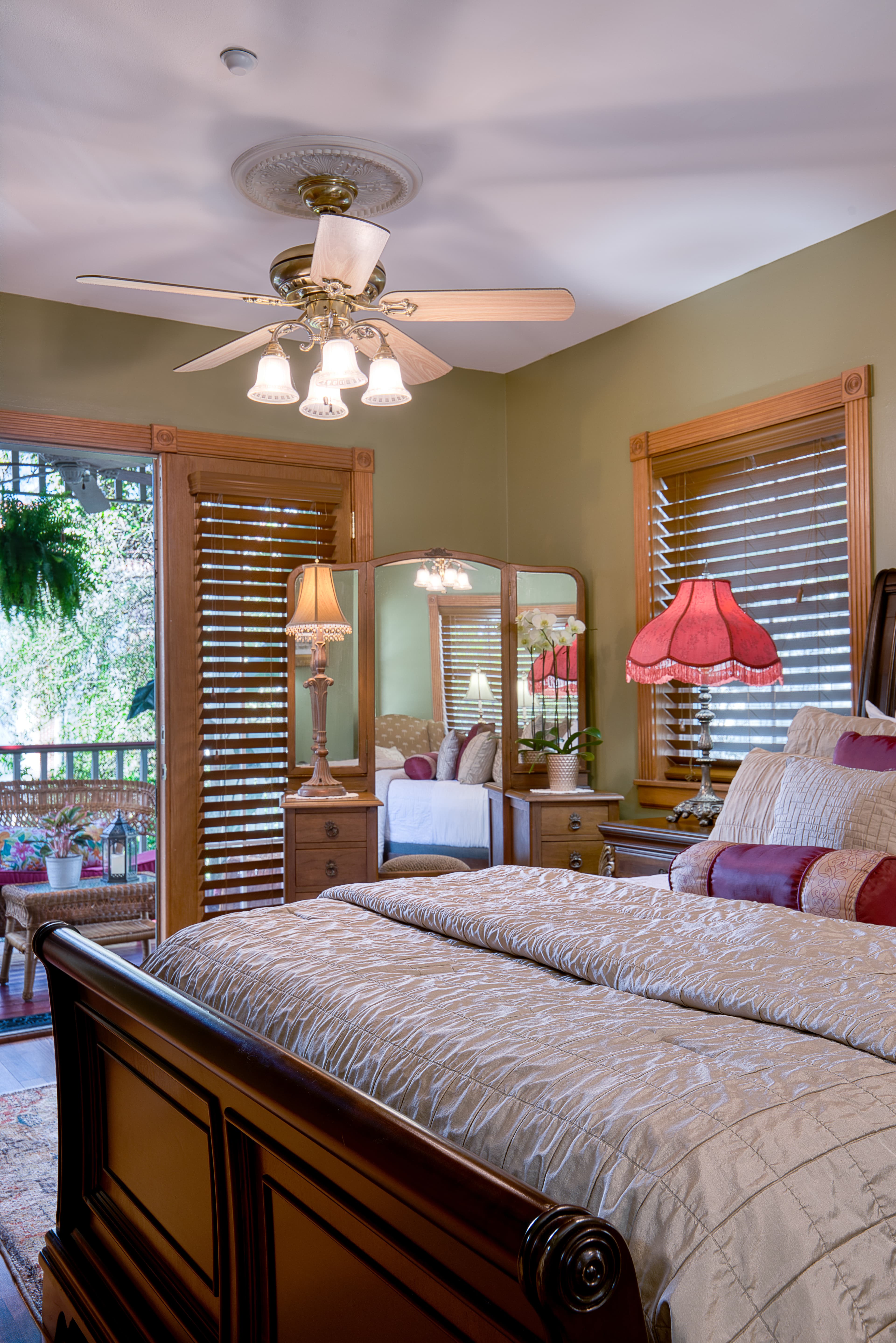 A traditionally styled bedroom with a dark wood bed frame, a sitting area with a red lamp, and a balcony with a view through a glass door.