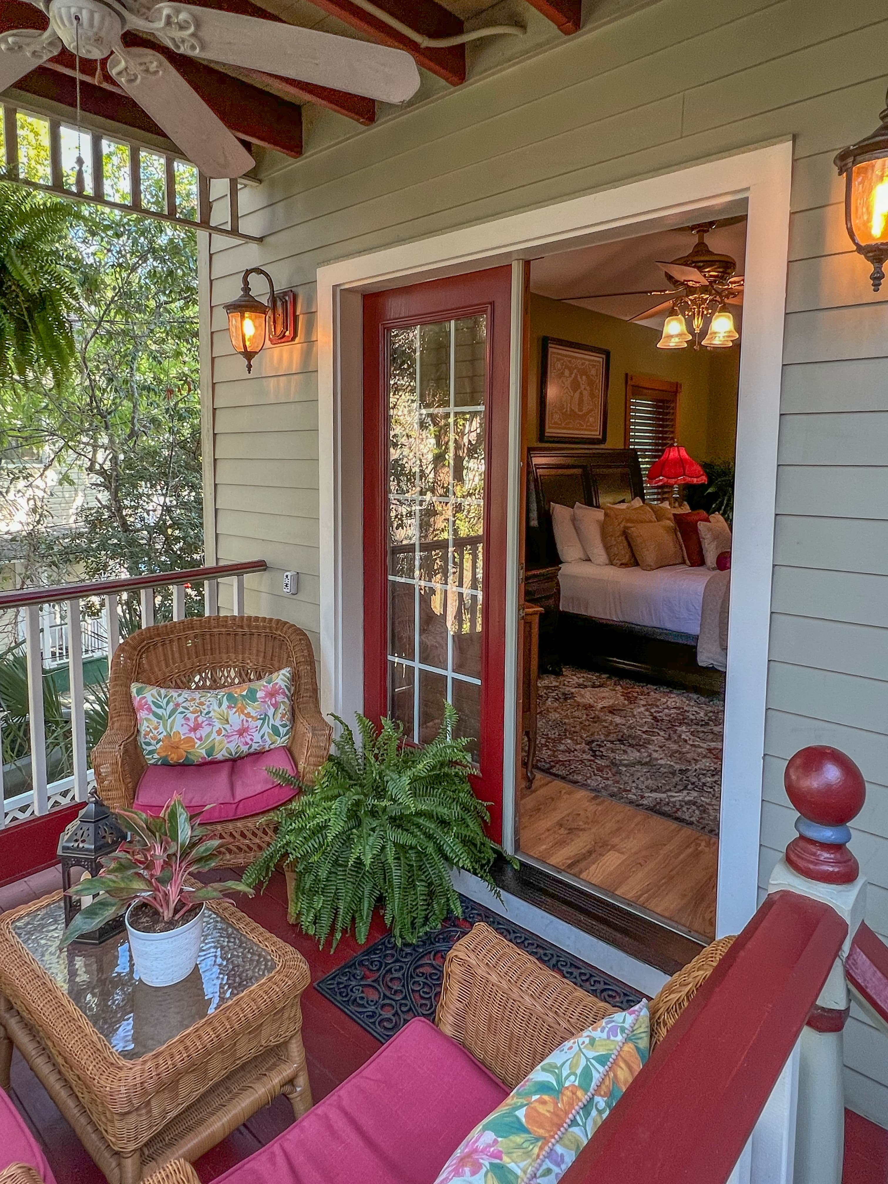 A screened-in porch with wicker furniture and a red door leading to a bedroom with a bed and a lamp. The porch overlooks a lush green outdoor area.