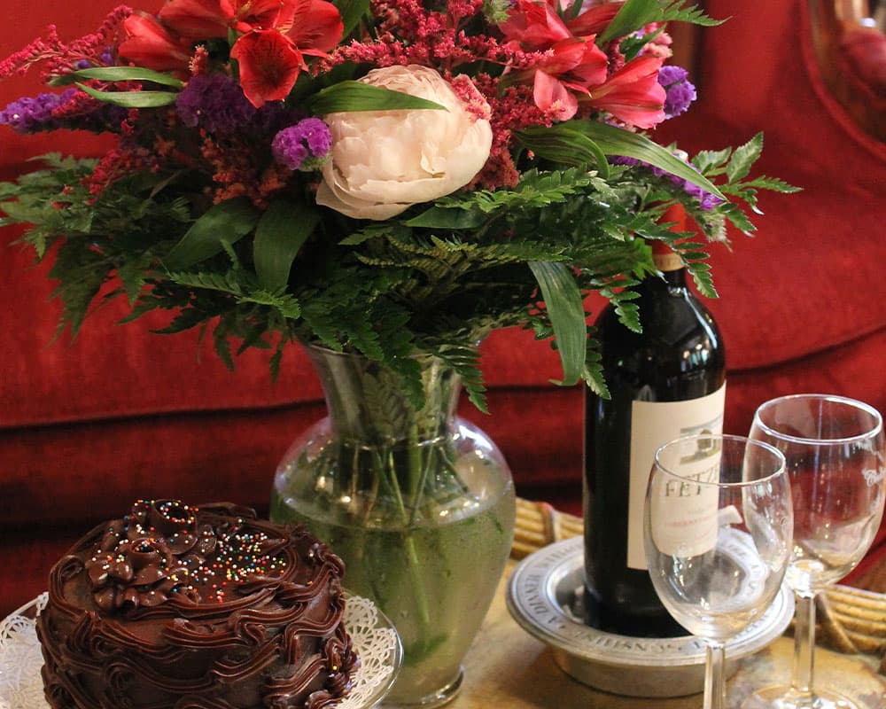 A festive arrangement featuring a vibrant floral bouquet, a bottle of wine, two glasses, and a chocolate cake.