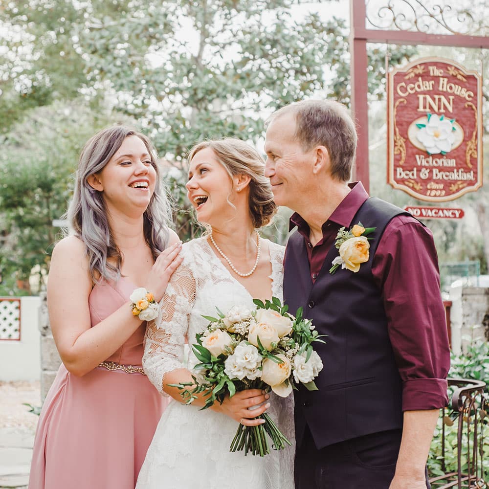 A smiling bride stands between her two joyful companions outside a charming inn, holding a bouquet of flowers.