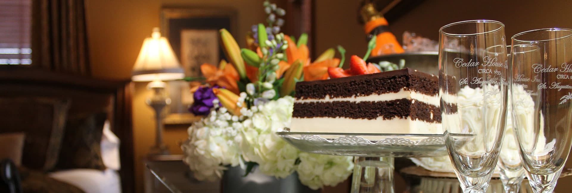 A chocolate layer cake is displayed with elegant floral arrangements and champagne flutes in a cozy room setting.