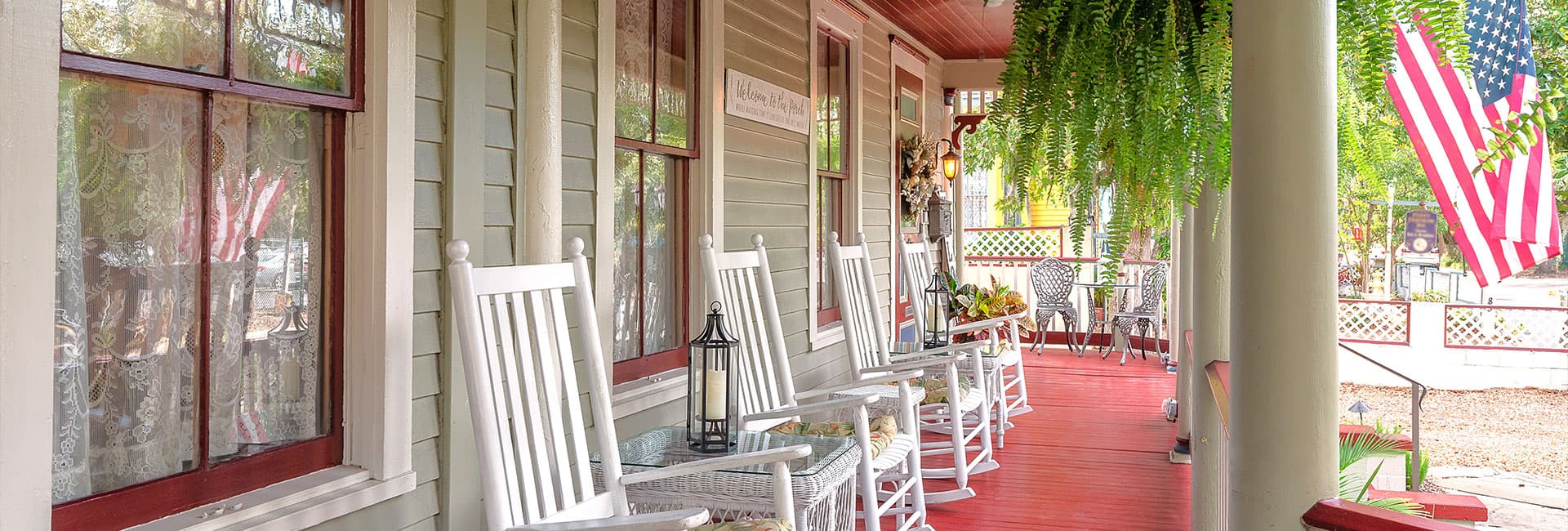 A charming porch with rocking chairs, a lantern, and hanging ferns, featuring a view of an American flag in the background.